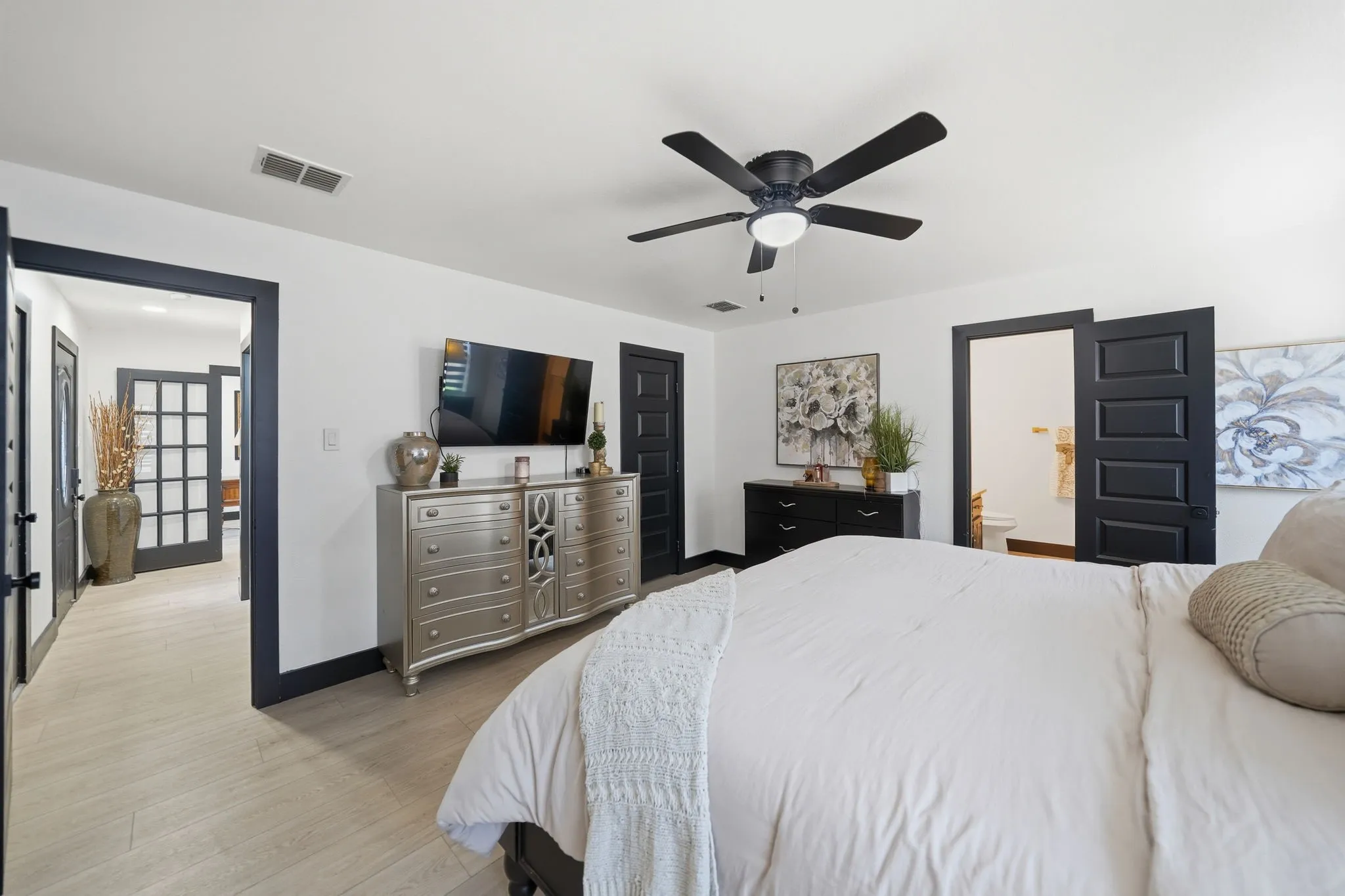 Spacious bedroom featuring light wood-finish flooring, a ceiling fan with integrated lighting, and white walls complemented by dark trim and doors