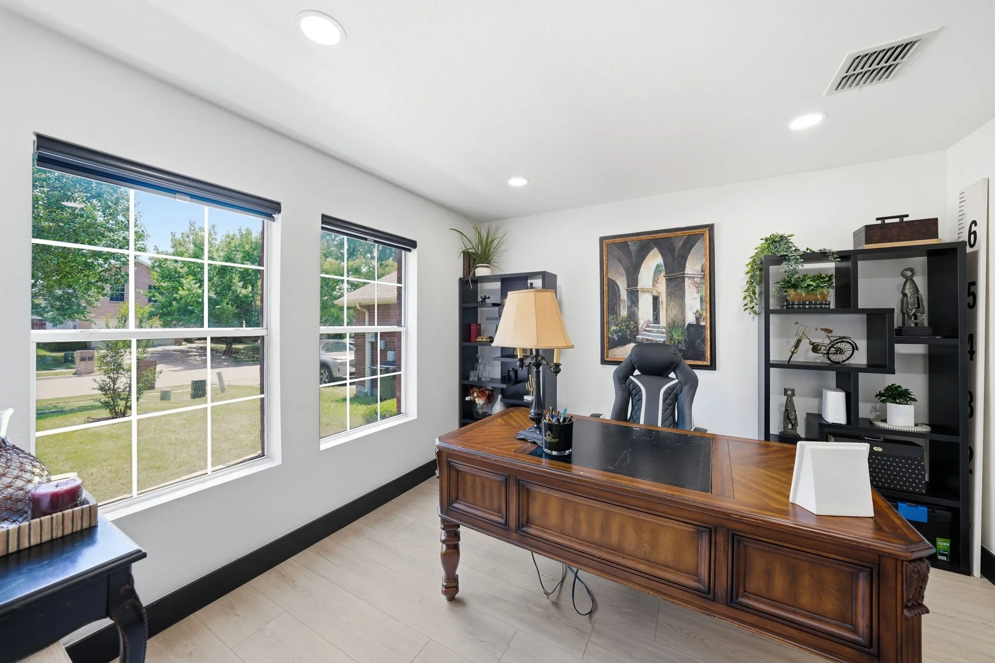 Bright interior space featuring two large windows with white grids, recessed lighting, wood-finish flooring, and a dark wood executive desk
