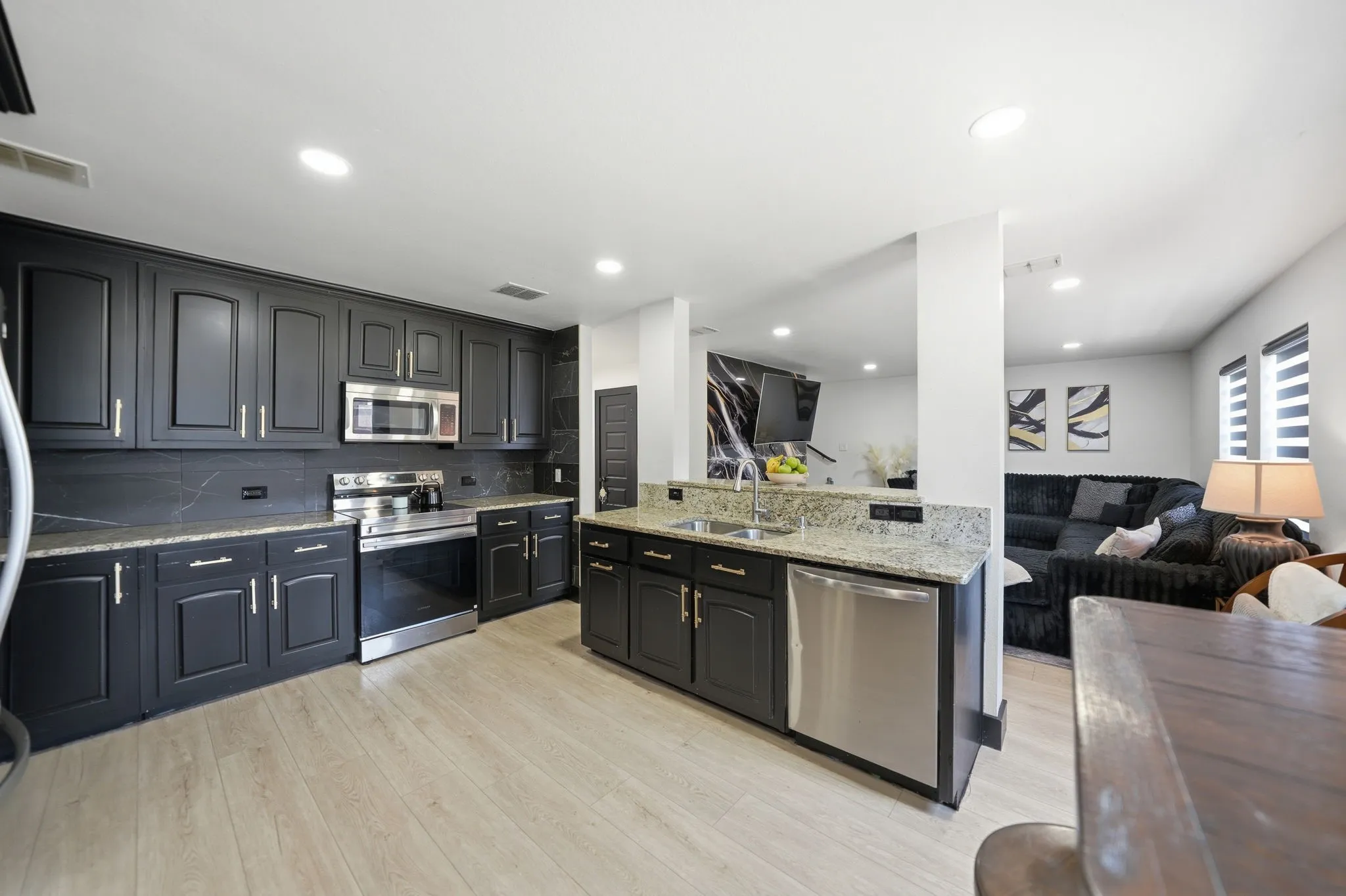 Modern kitchen featuring dark cabinetry with gold-toned hardware, stainless steel appliances, speckled stone countertops, and light wood-finish flooring