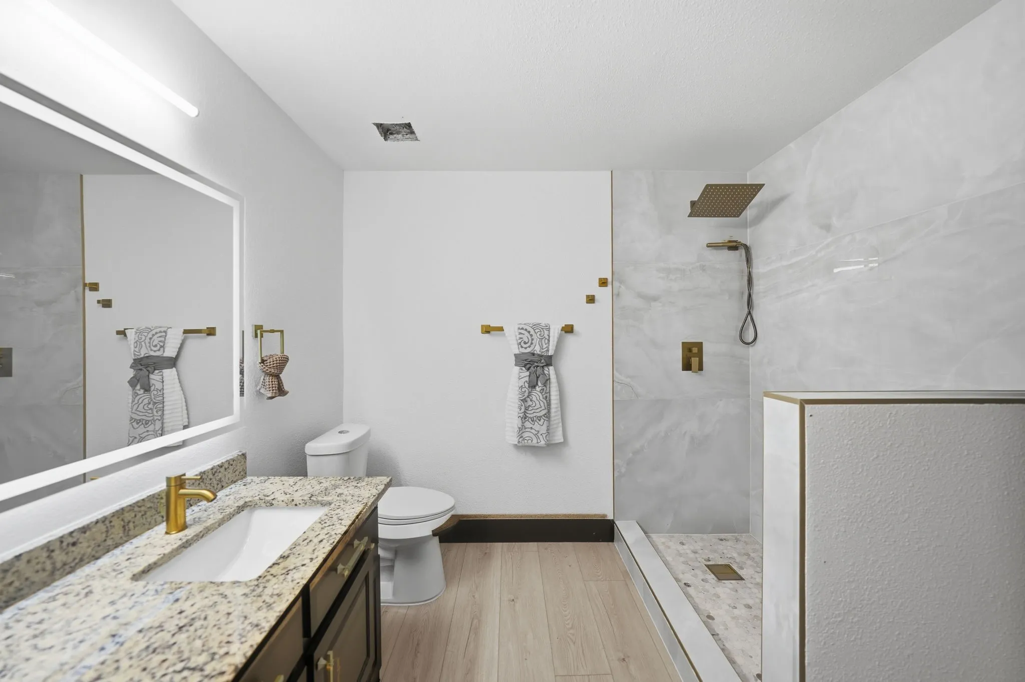 Modern bathroom featuring a walk-in shower with a rain showerhead, a single vanity with a stone countertop and gold-finish fixtures, a lighted mirror, and light wood-finish flooring