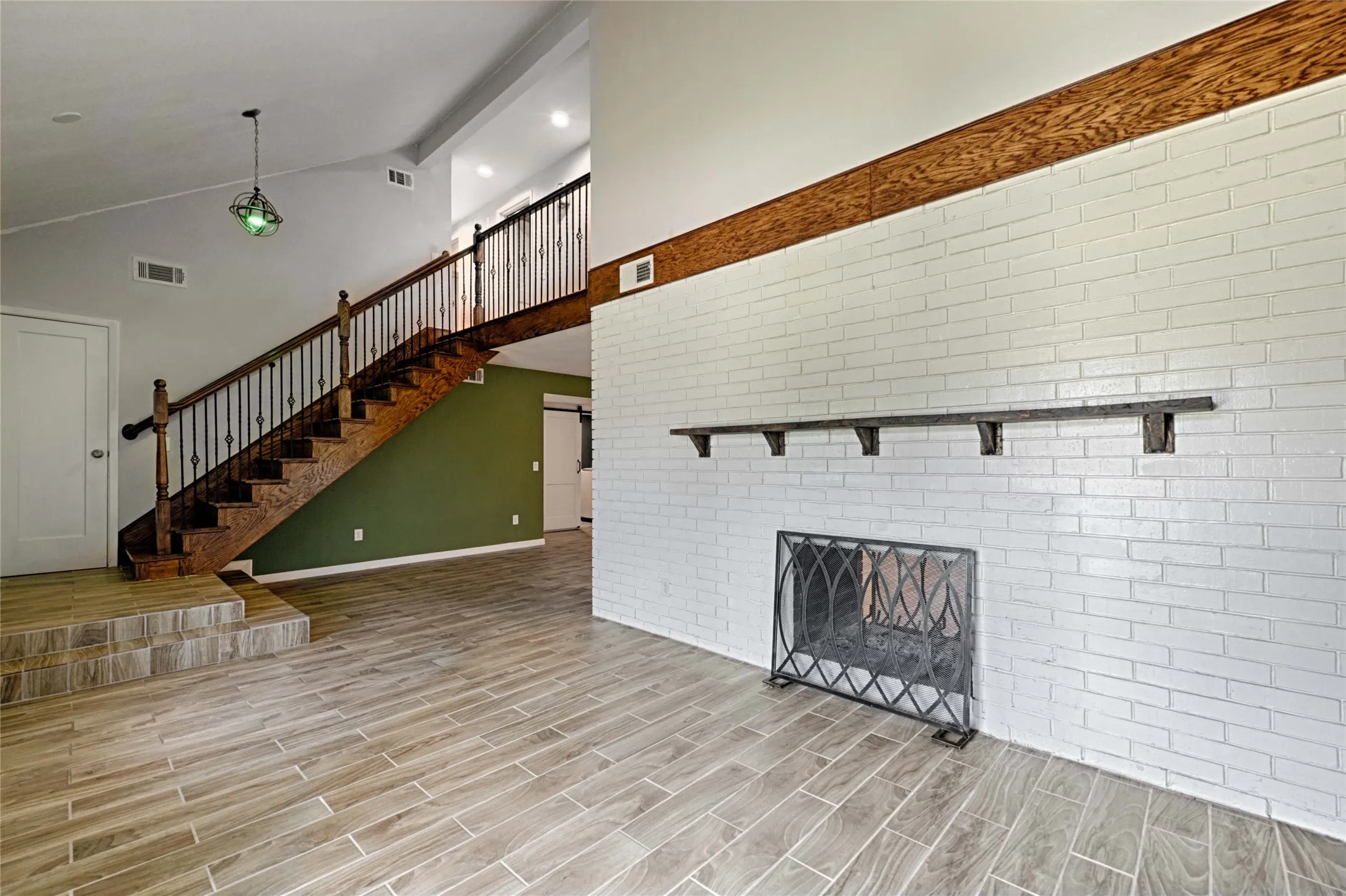 Open living area featuring wood-finish tile flooring, a white-painted brick fireplace with a decorative screen, a wood mantel, and a dark wood staircase with wrought iron balusters