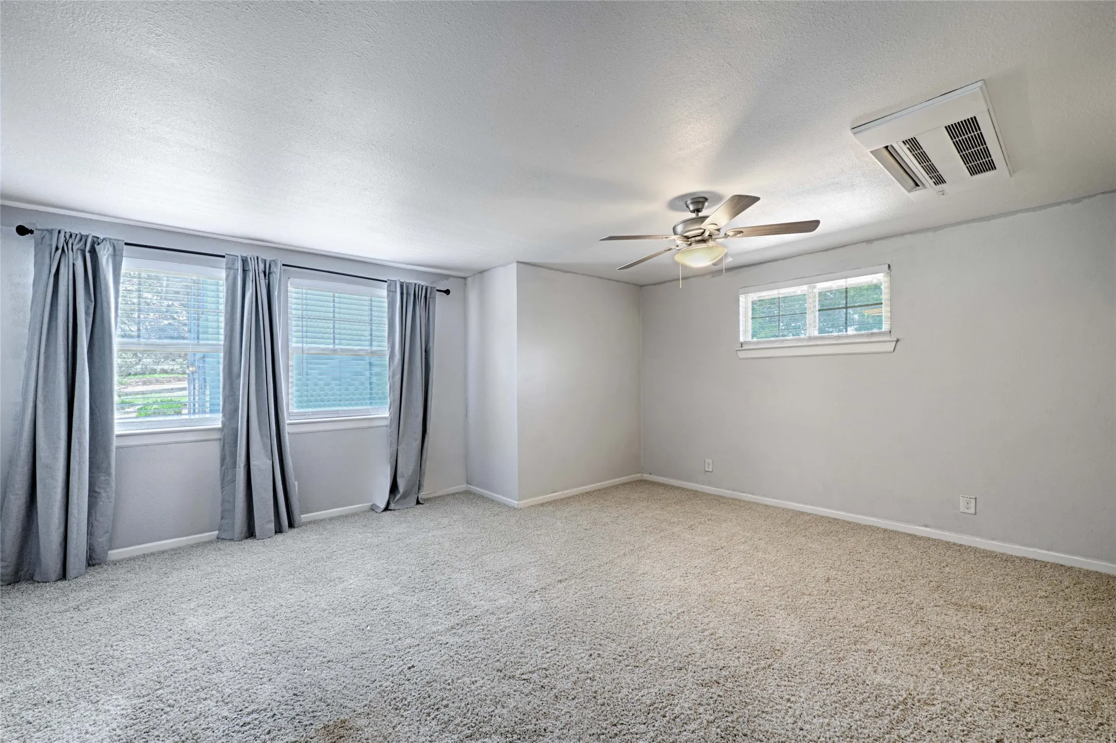 Spacious room featuring light-toned carpeting, two large windows with blinds and drapery, and a ceiling fan with integrated lighting