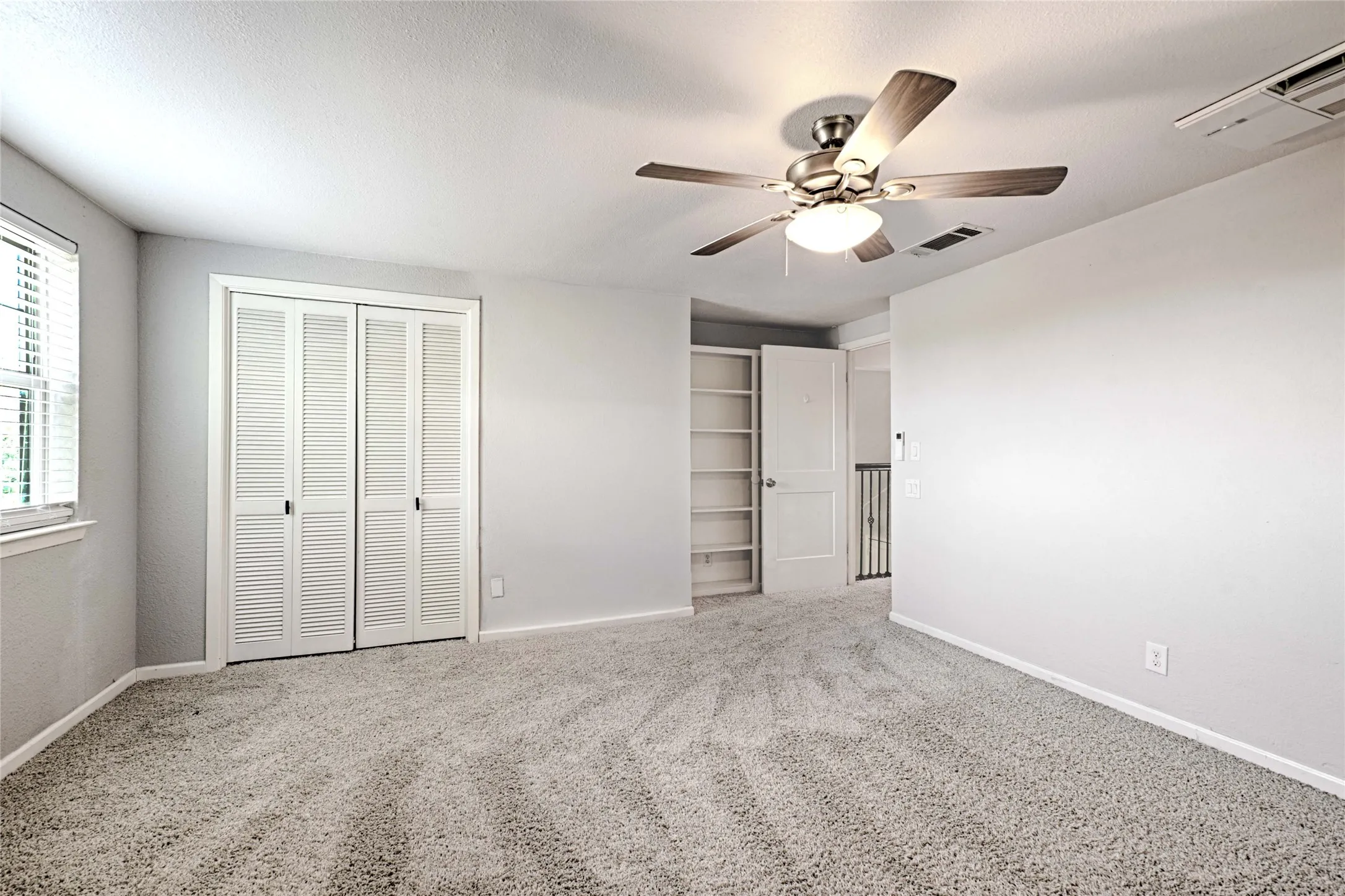 Spacious room featuring neutral carpet flooring, a ceiling fan with integrated lighting, window with blinds, louvered bi-fold closet doors, and built-in shelving