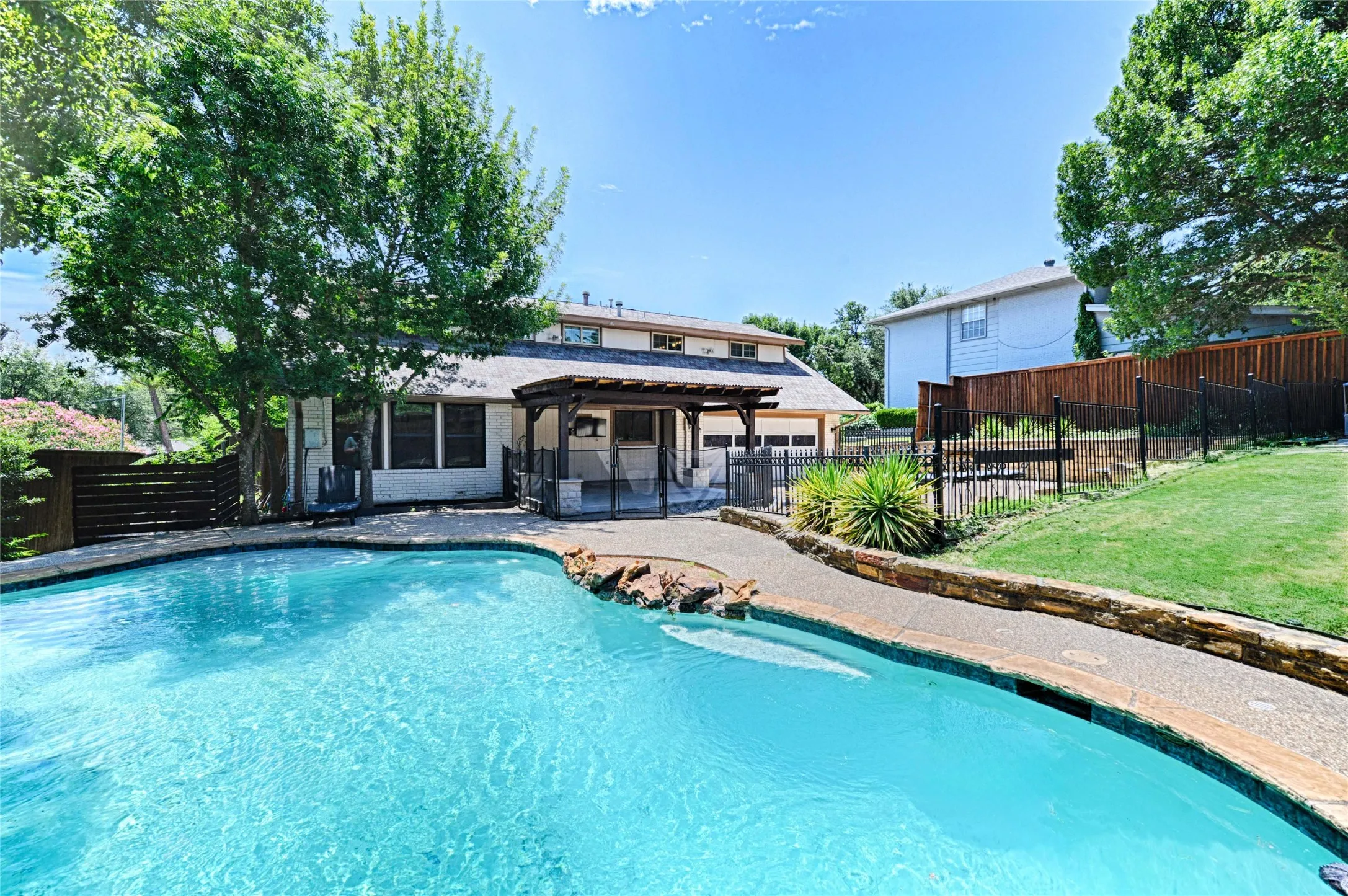 Backyard featuring a freeform swimming pool with rock accents, a covered patio area, and a tiered lawn with fencing