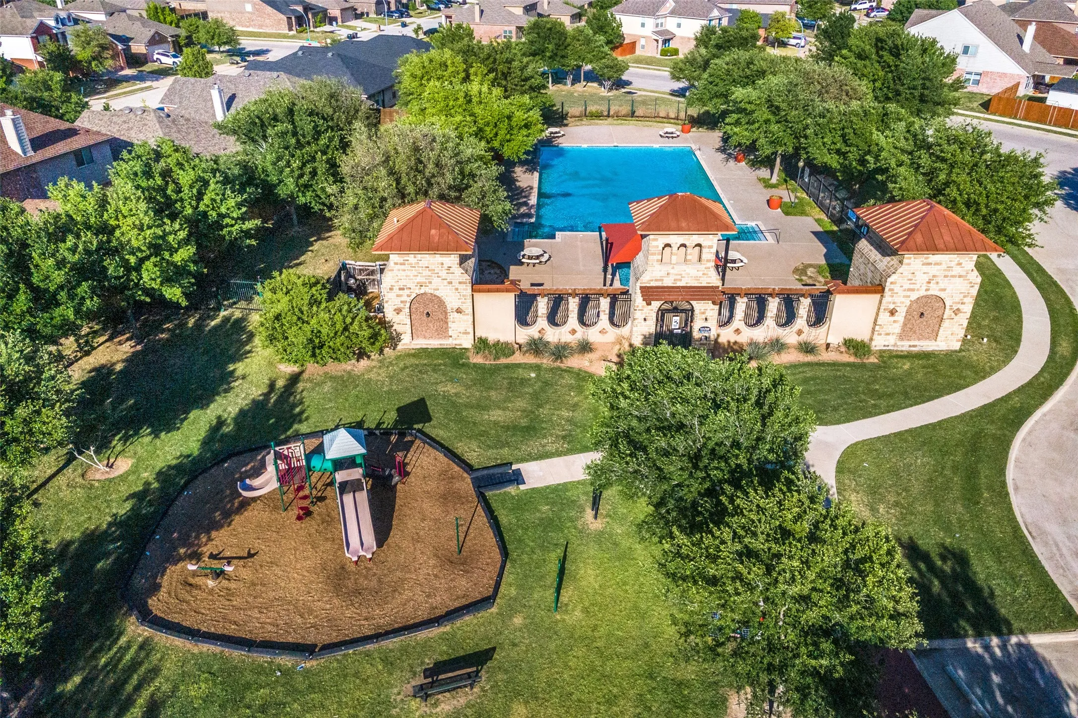 Community clubhouse with stone veneer and terracotta-style roof, featuring an outdoor swimming pool, adjacent playground with wood chip surfacing, and winding paved pathways surrounded by manicured green spaces