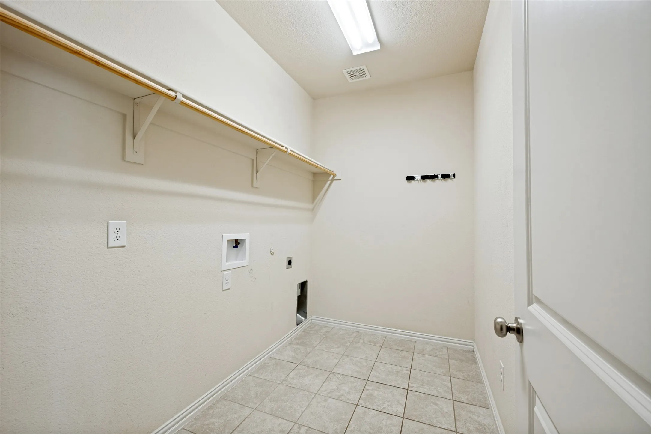 Utility area featuring light-toned tile flooring, overhead shelving, a clothing rod, and a flush-mount fluorescent light fixture