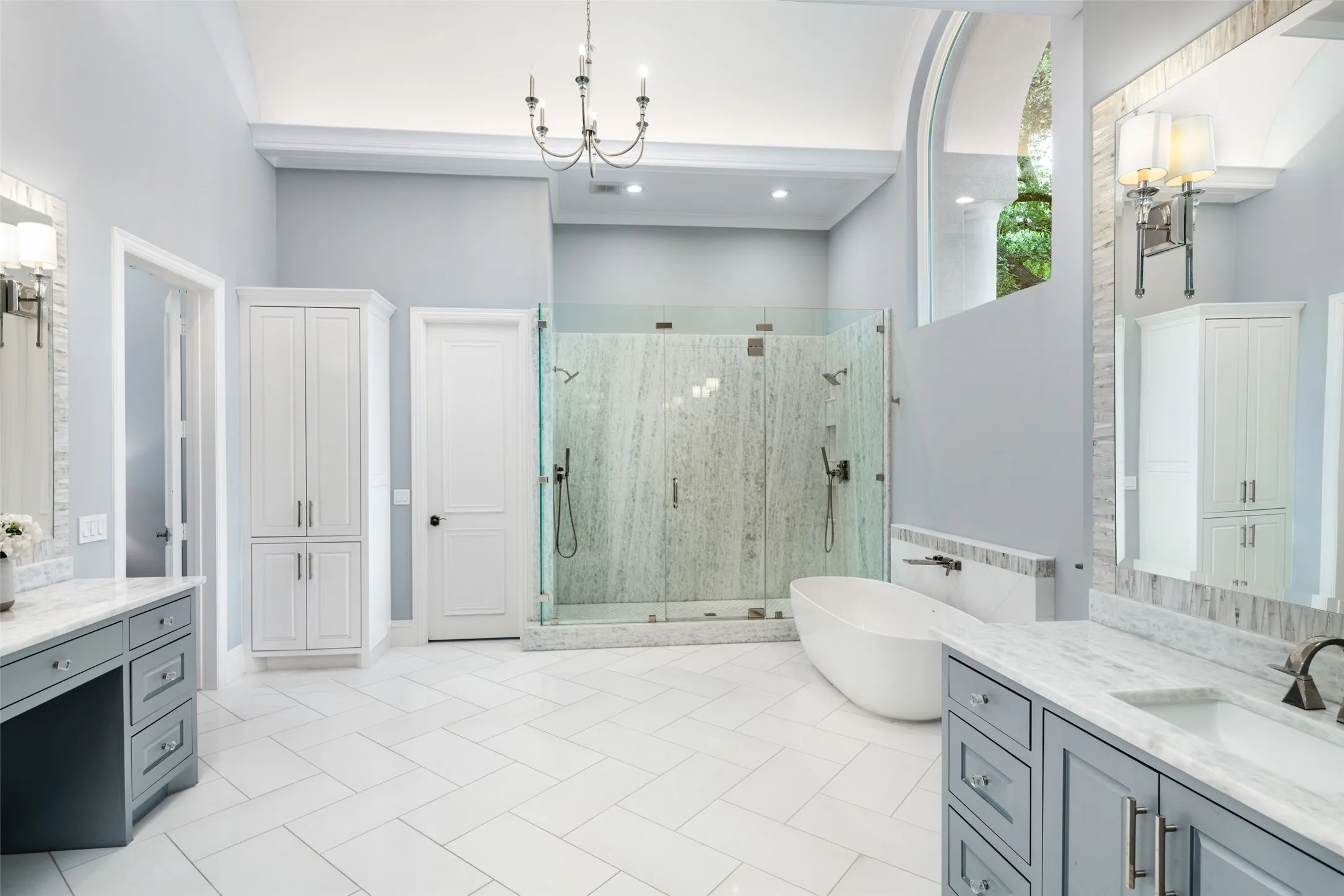 Spa-inspired primary bath offers dual vanities, freestanding soaking tub, oversized marble shower with dual rain heads, soaring ceilings, and abundant natural light from dramatic arched windows.