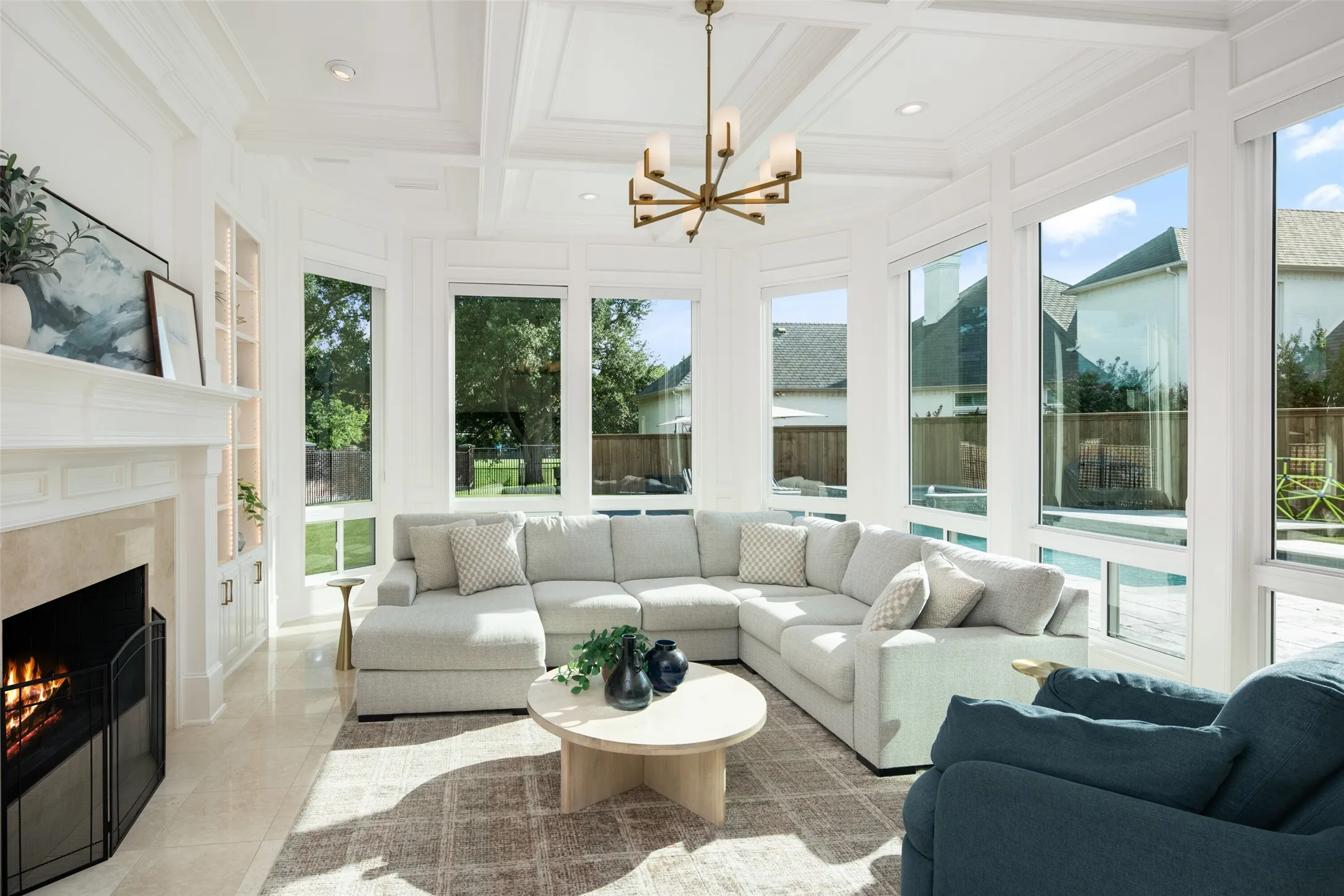 Walls of windows frame beautiful backyard and pool views while the coffered ceiling, fireplace, and spacious seating area create a warm, inviting atmosphere filled with natural light.