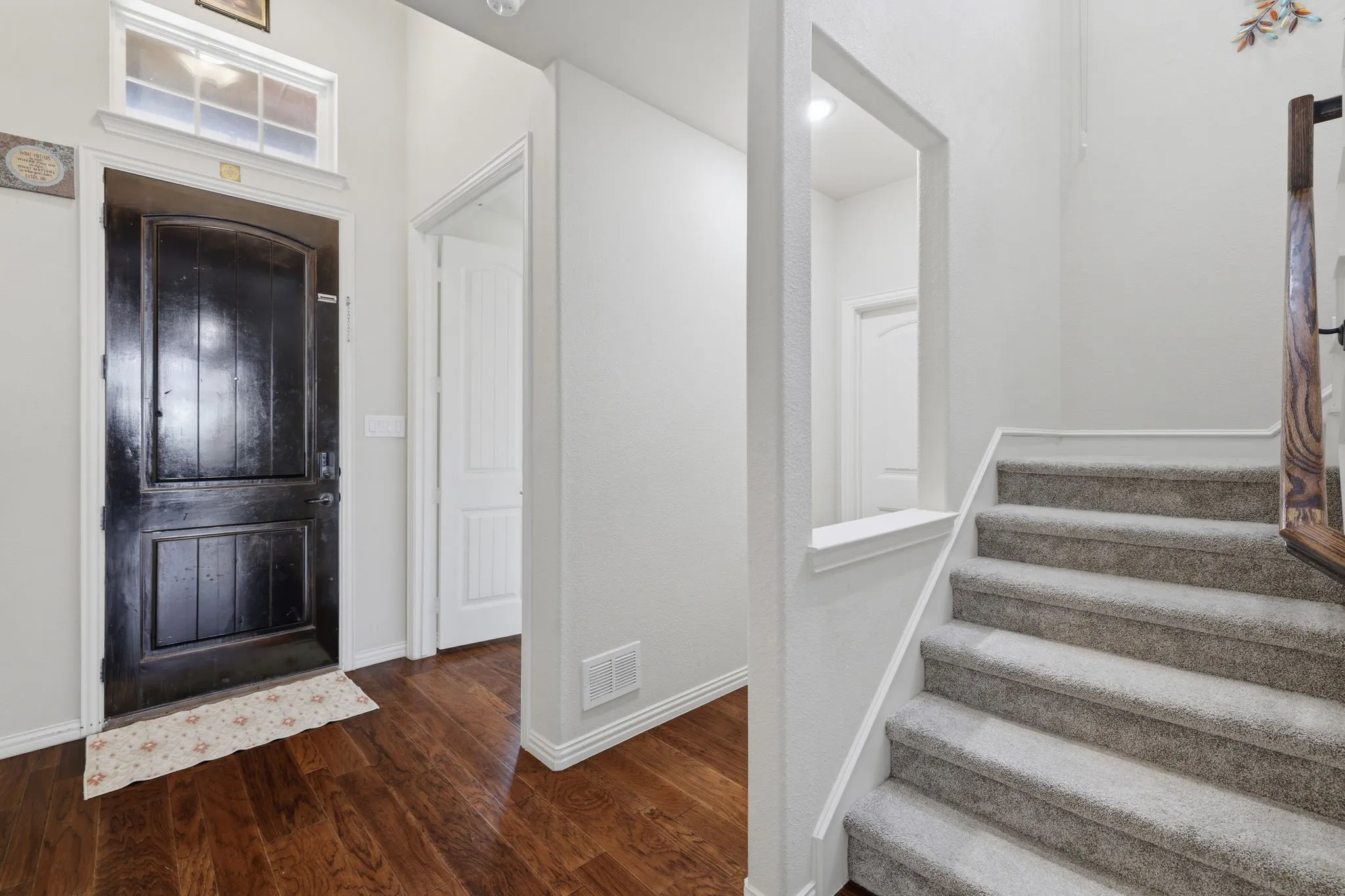 Entryway featuring a dark wood-finish door with an arched top panel and transom window, wood-finish flooring, white baseboards, a carpeted staircase with wood handrail, and a framed wall opening