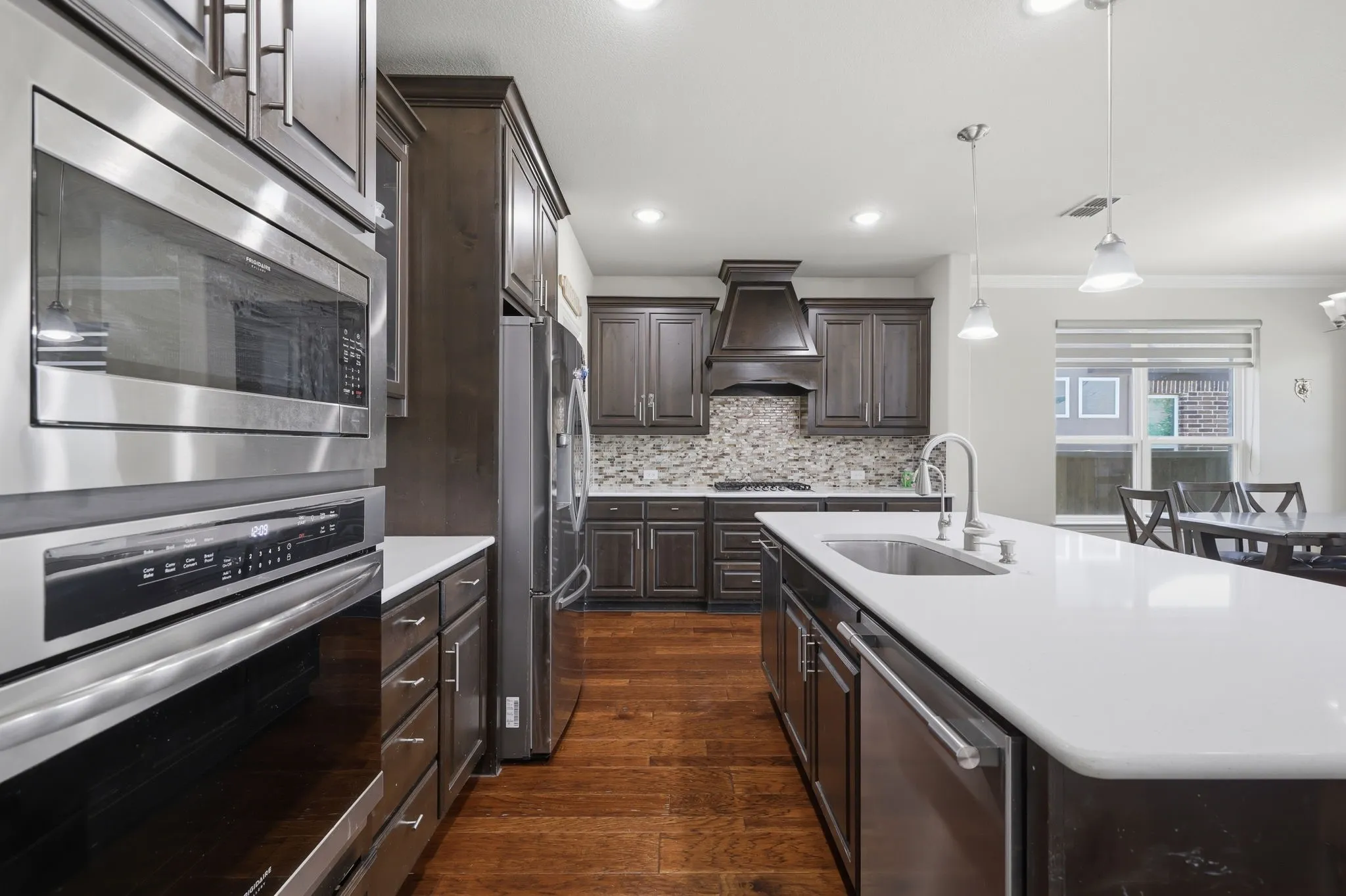 Contemporary kitchen featuring stainless steel appliances, dark wood-finish cabinetry, a built-in microwave and oven, a center island with a sink, and pendant lighting