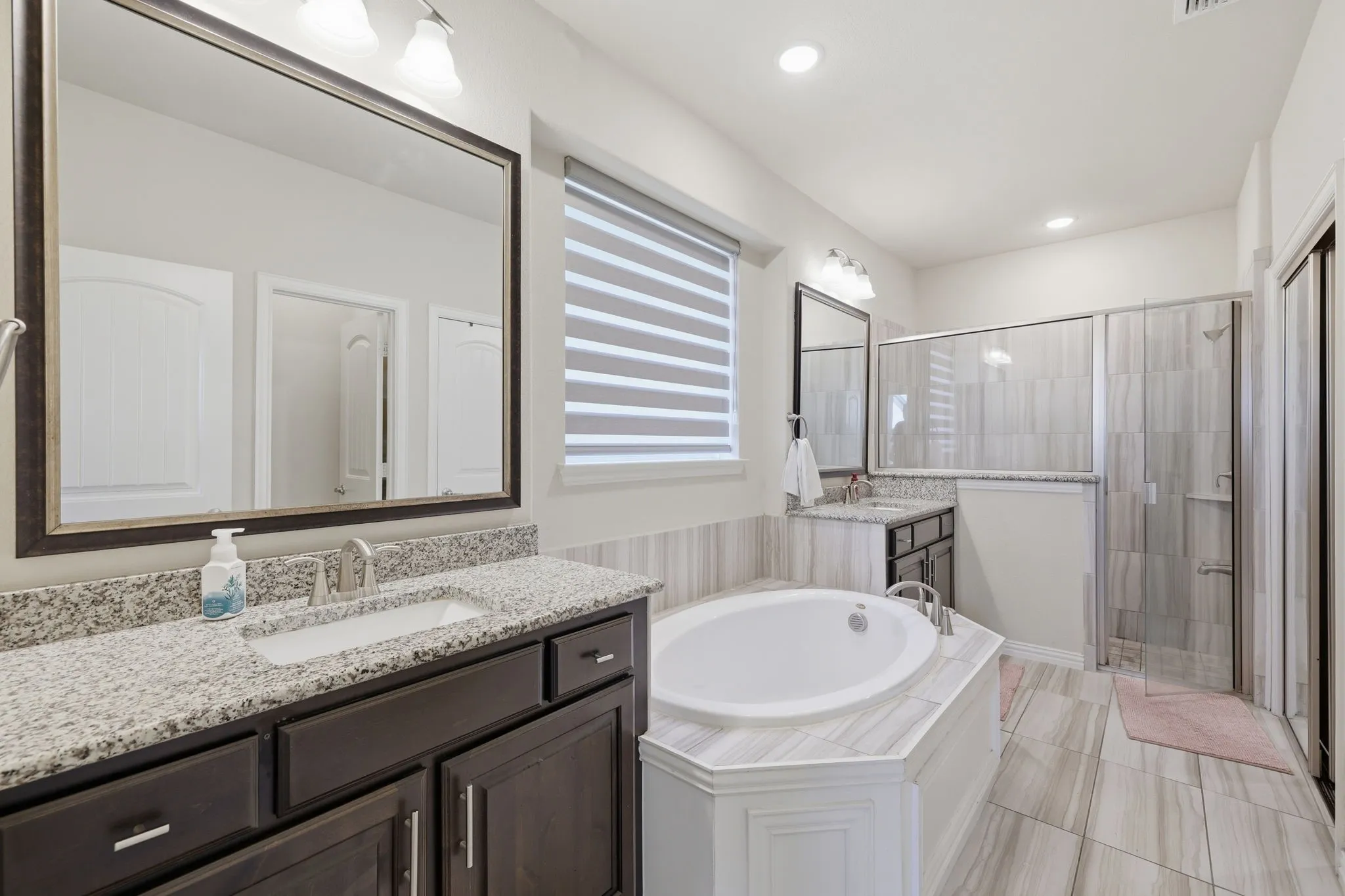 Spacious bathroom featuring a built-in soaking tub, separate glass-enclosed shower, dual vanity with stone countertops, tiled flooring, and a large framed mirror