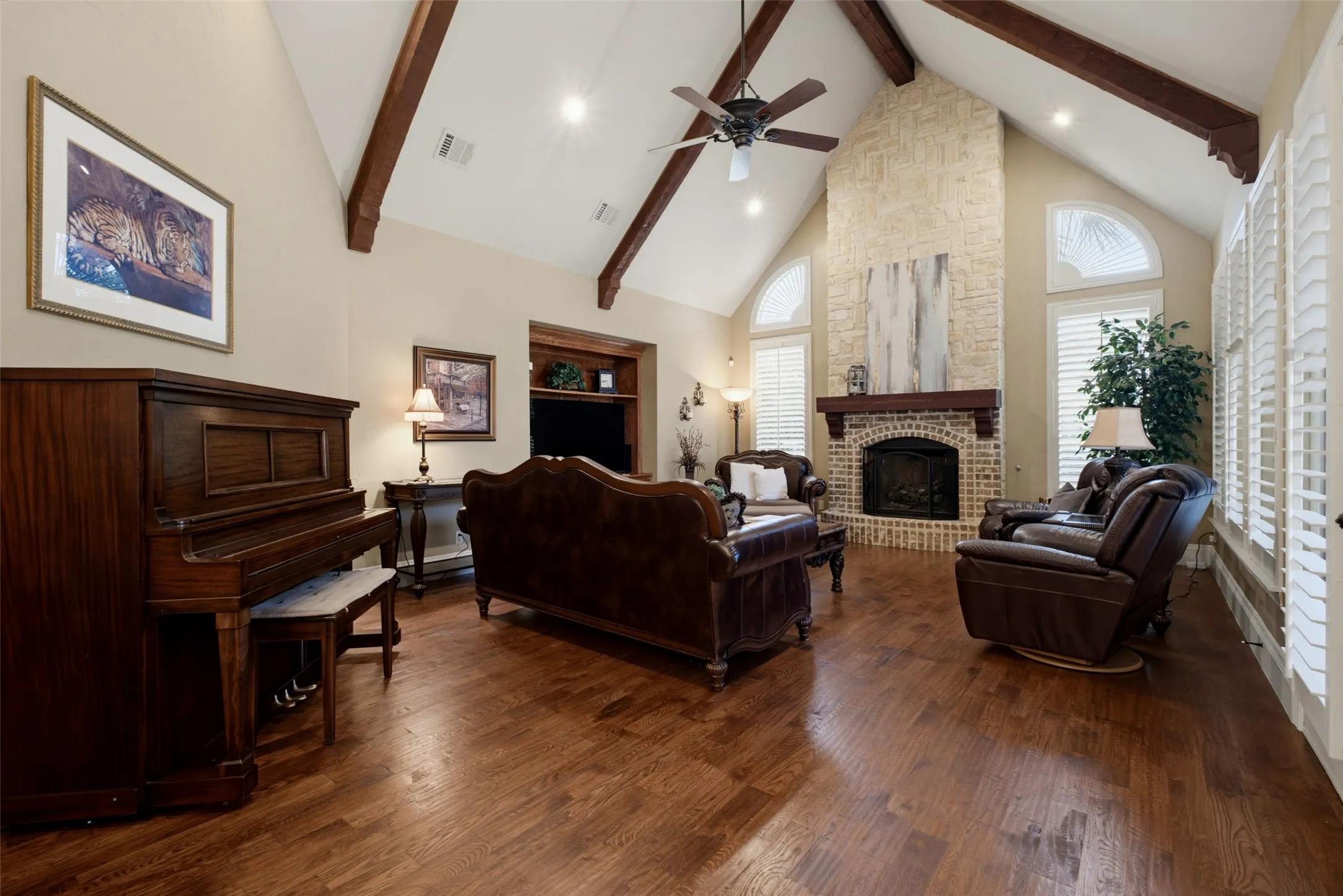 Vaulted ceilings with exposed wood beams, hardwood flooring, stone fireplace with a wood mantle, multiple tall windows with plantation shutters, and a built-in media niche