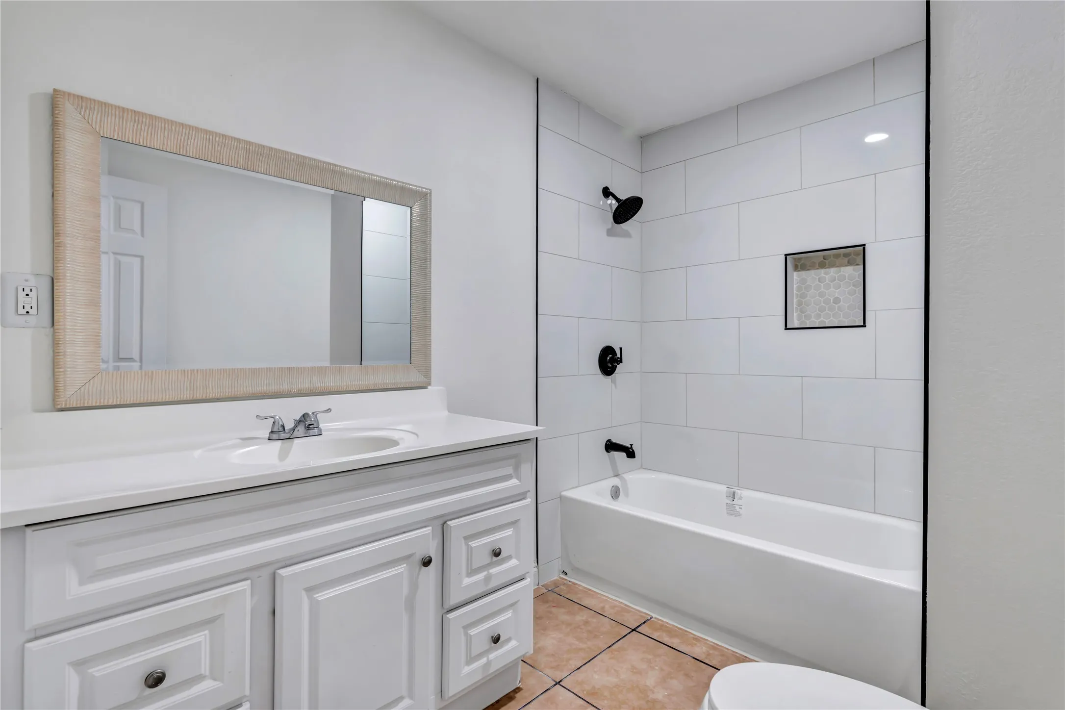 Bathroom featuring a white vanity with a single basin, a framed mirror, a bathtub with white subway tile surround, a recessed shower niche with patterned tile, and black plumbing fixtures