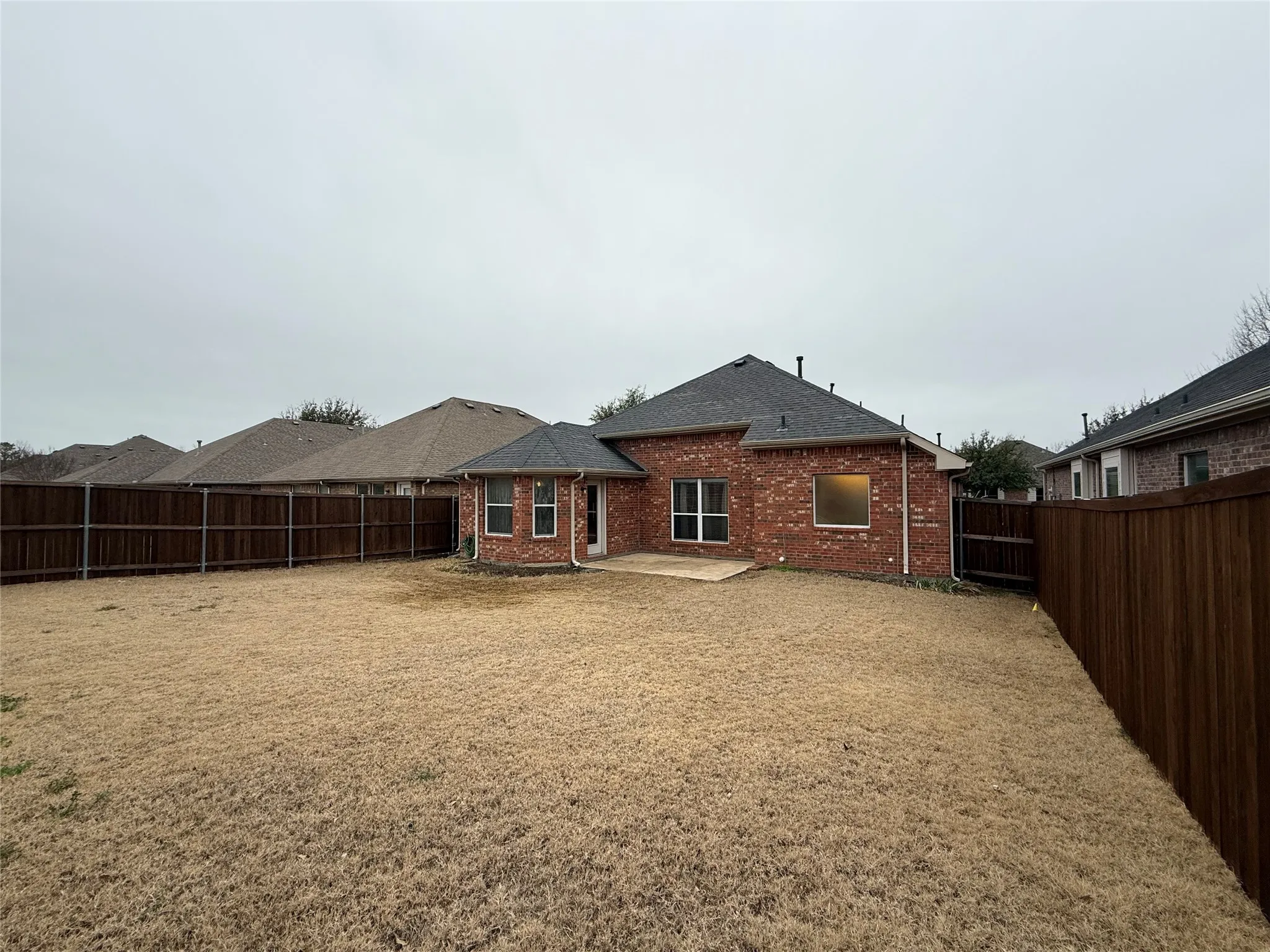 Expansive backyard featuring a brick exterior, a concrete patio, a dark-stained wood fence, and a dark shingled roof