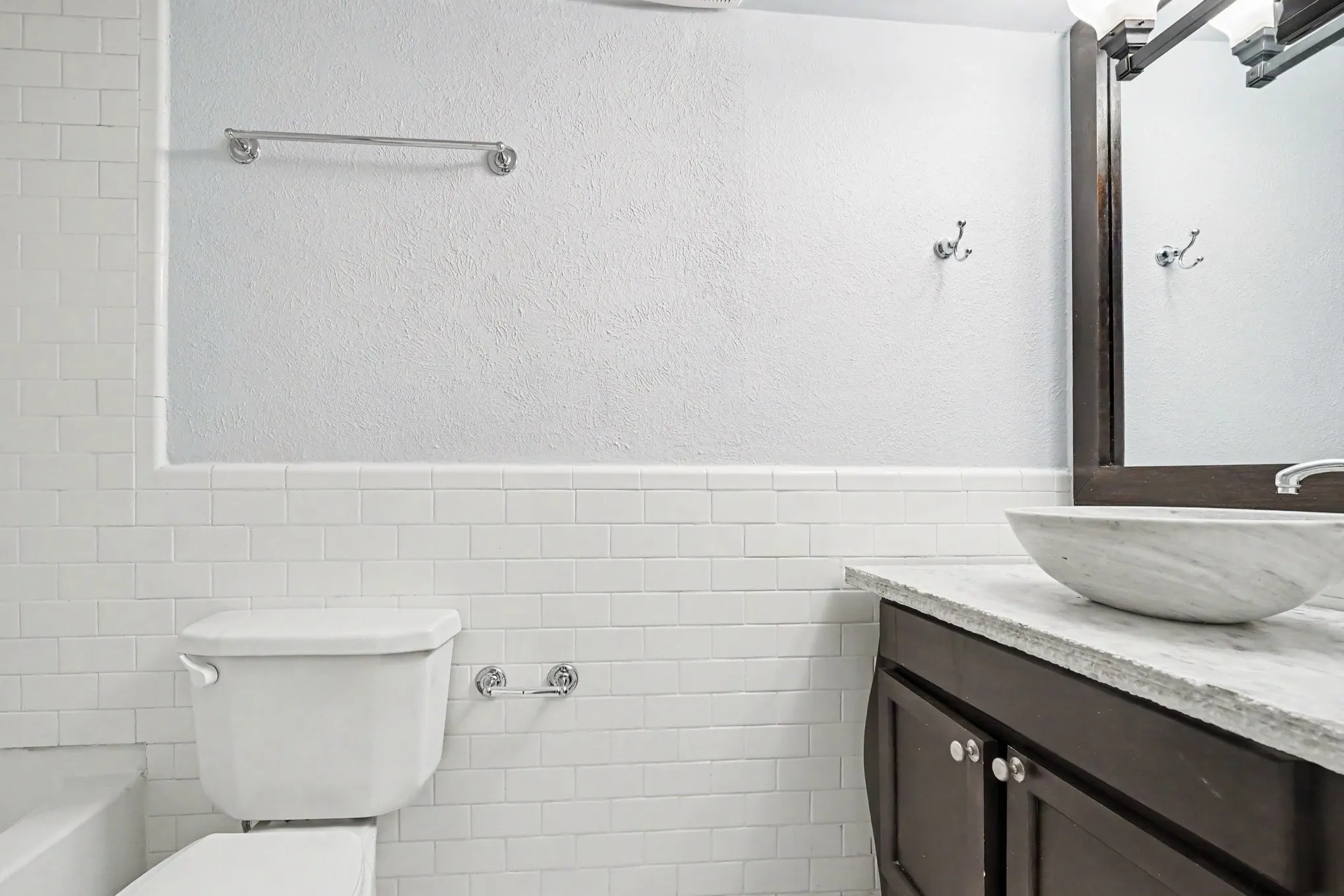 The spacious hall bathroom features a vanity with a vessel sink, stone countertop, dark wood-finish cabinetry, and a framed mirror.  A tub with a shower is to the left in this view.