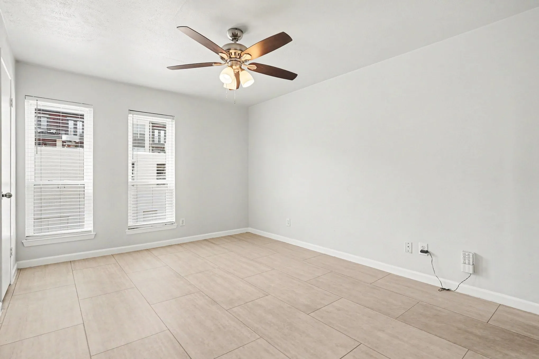 The primary bedroom features light-toned tile flooring, light gray walls, two vertical windows with blinds, a ceiling fan, a walk-in closet, and an ensuite bath.
