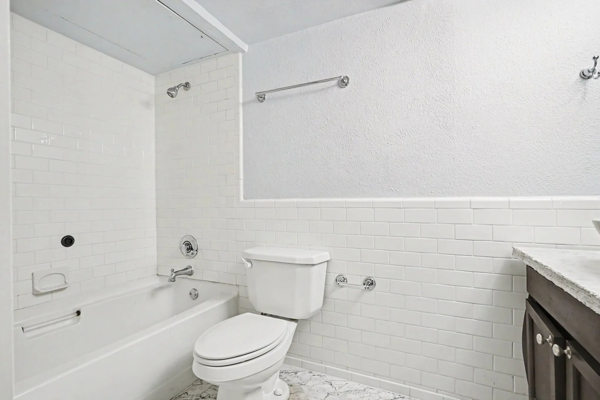 The spacious hall bath features white subway tile tub surround, white bathtub, chrome shower fixture, and an extended seat toilet.  The vanity is to the right in this view.