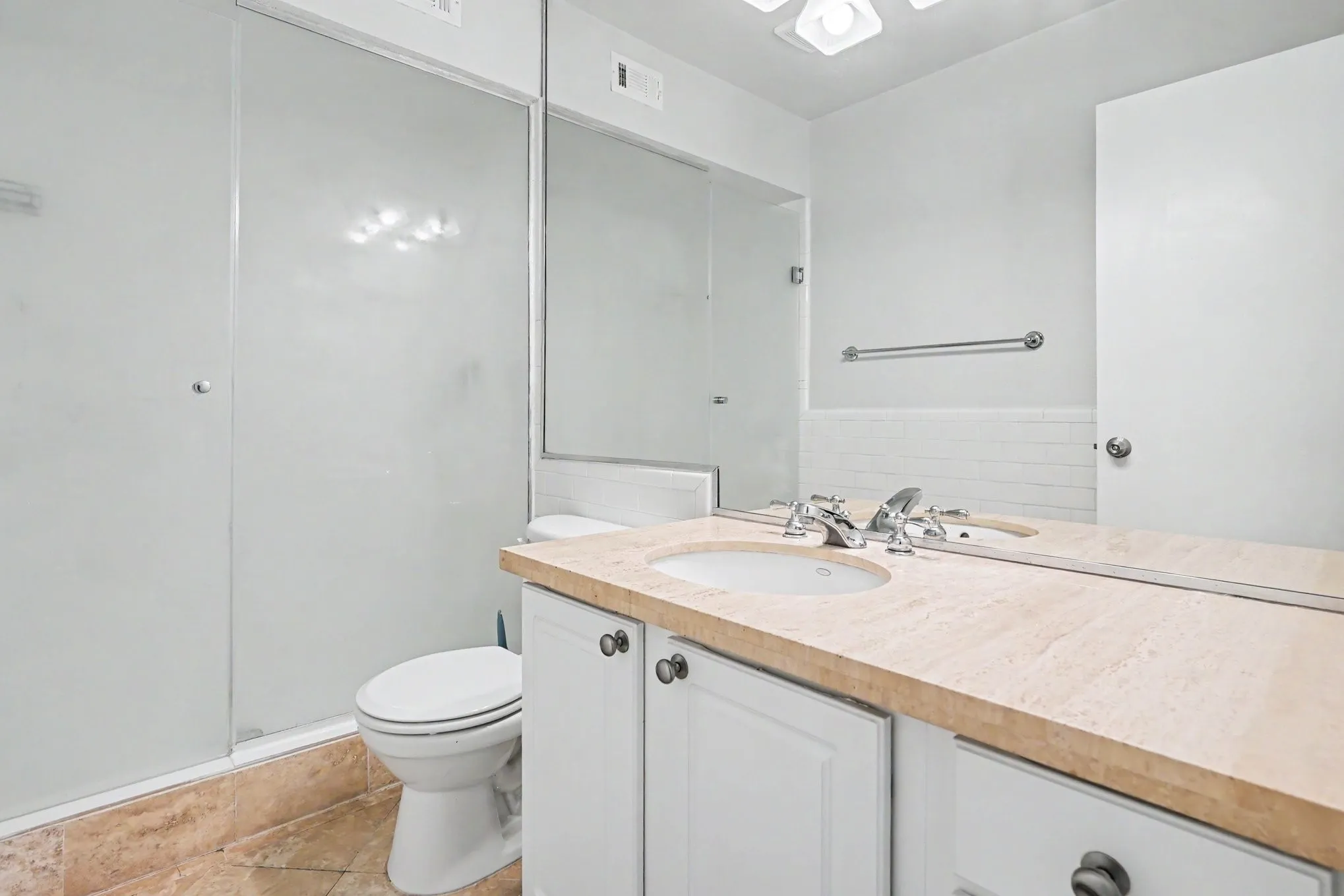 The primary bedroom ensuite bathroom features a single vanity with a light-toned countertop, an integrated sink, travertine flooring, a frosted glass shower with white subway tile, and white cabinetry