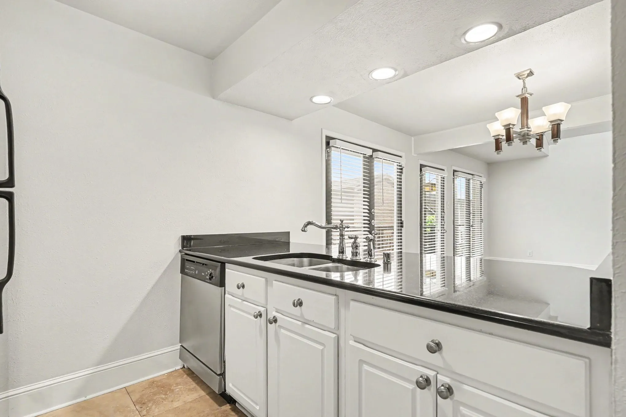 This kitchen view shows the stainless steel dishwasher, an under-mount sink with a gooseneck faucet, white cabinetry, dark granite countertops, and travertine flooring.