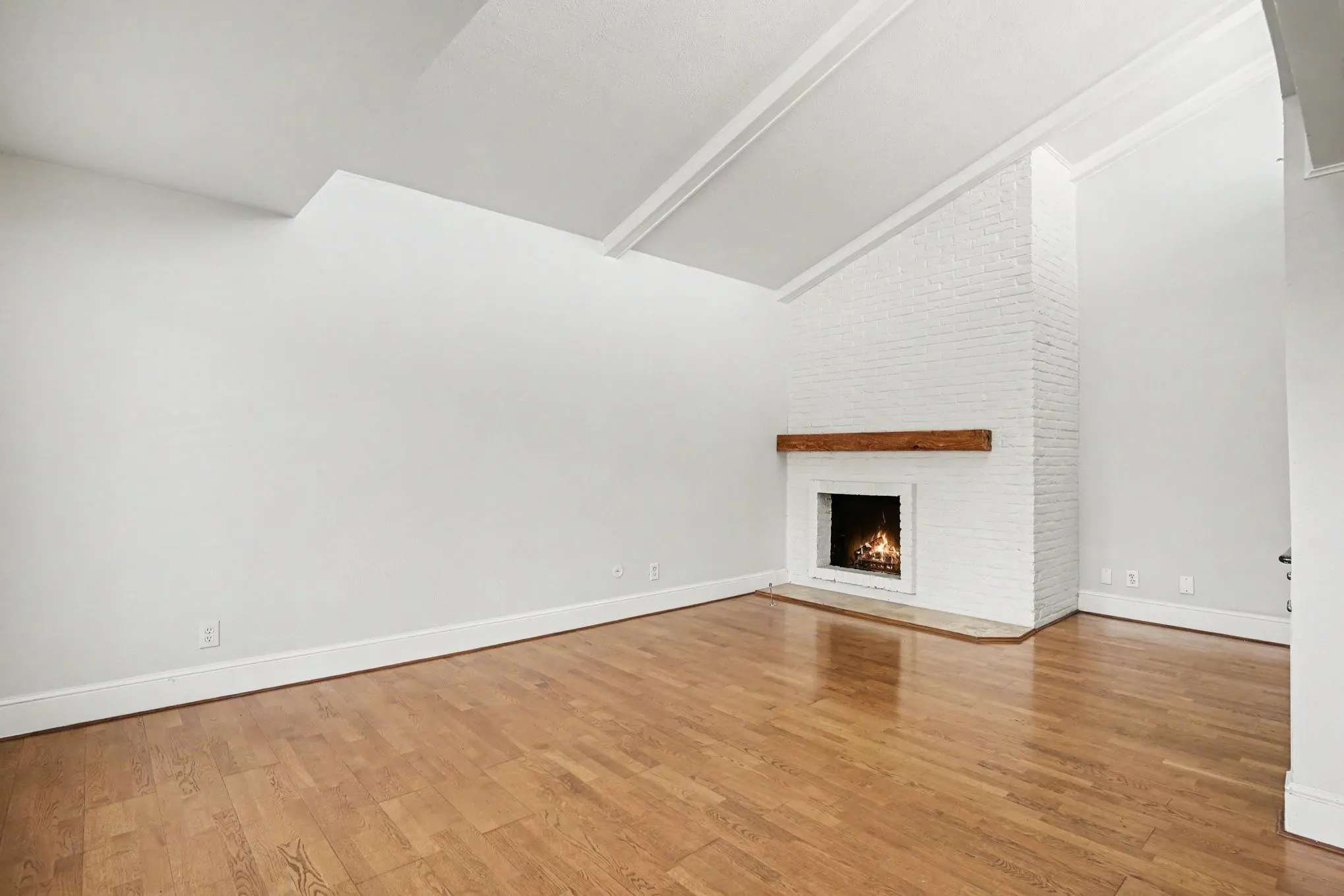 The spacious living room featuring a white painted brick fireplace with a wood mantel, vaulted ceiling with exposed beams, wood-finish flooring, and white baseboards. The entire unit was painted light gray in December 2025.