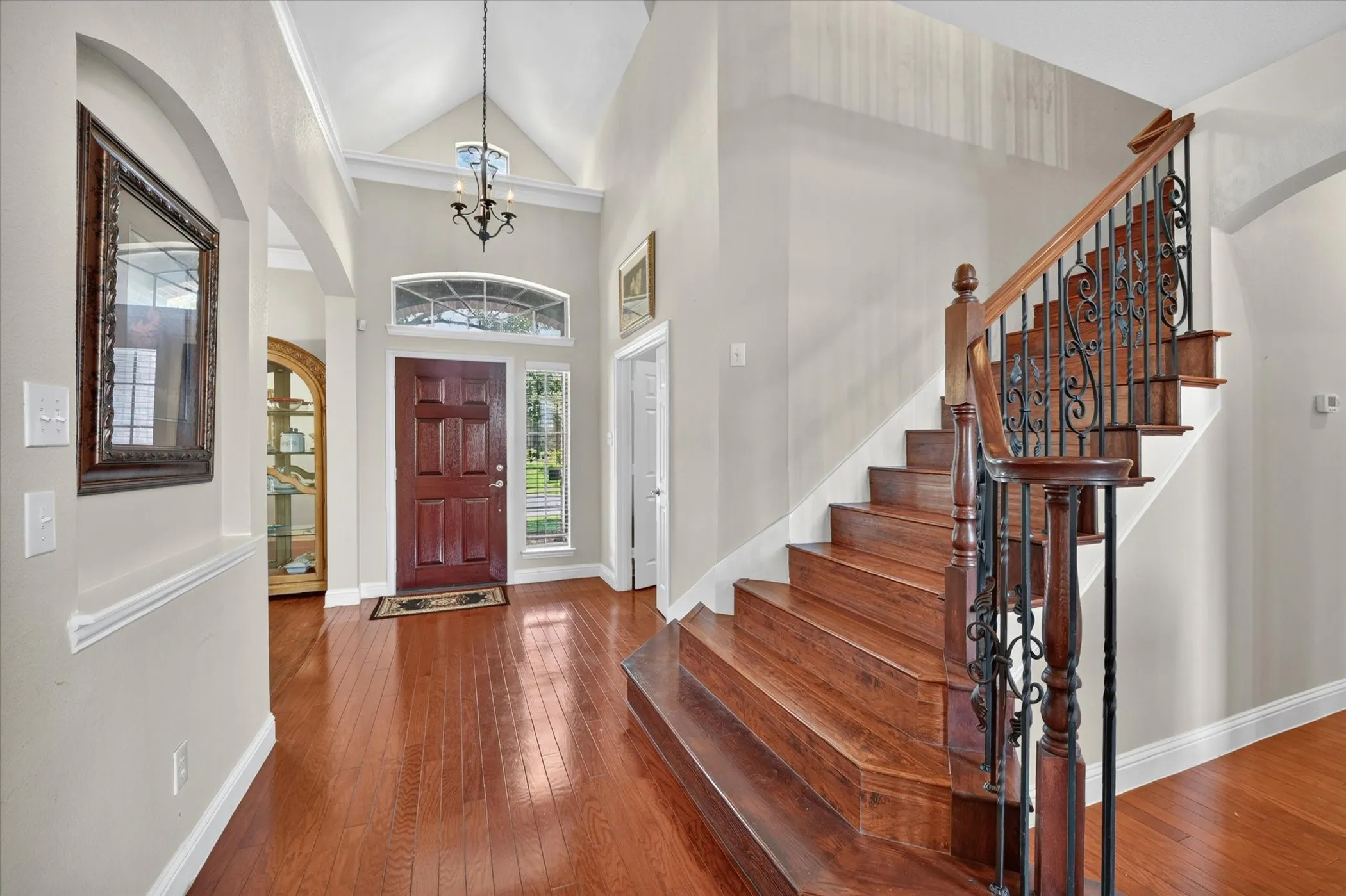 Grand foyer with soaring vaulted ceilings, hardwood flooring, a rich wood paneled entry door, arched transom window, and an elegant staircase featuring wrought iron balusters and a dark wood handrail