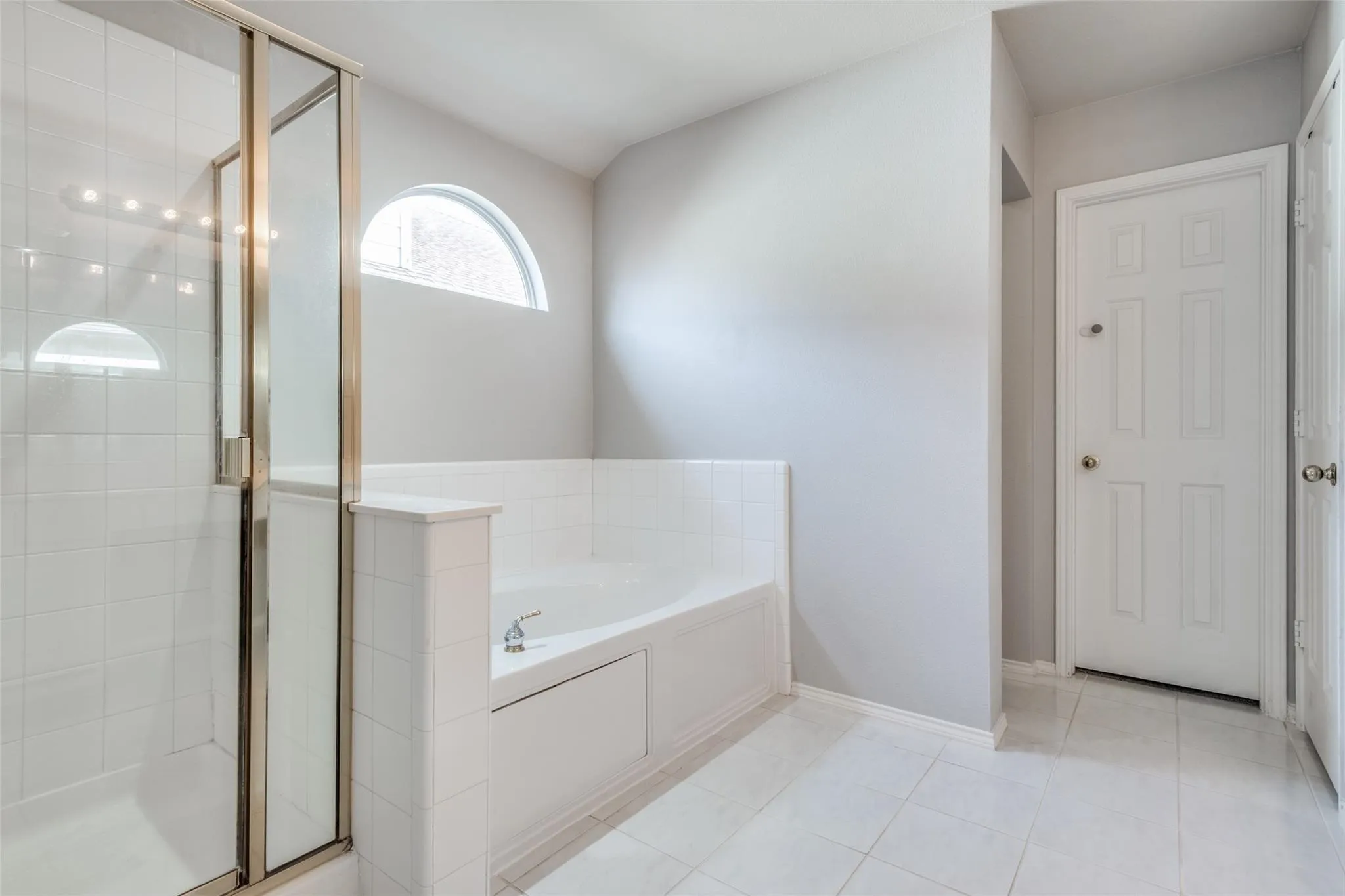Bathroom featuring a glass-enclosed shower, a built-in soaking tub, an arched transom window, and white tile flooring