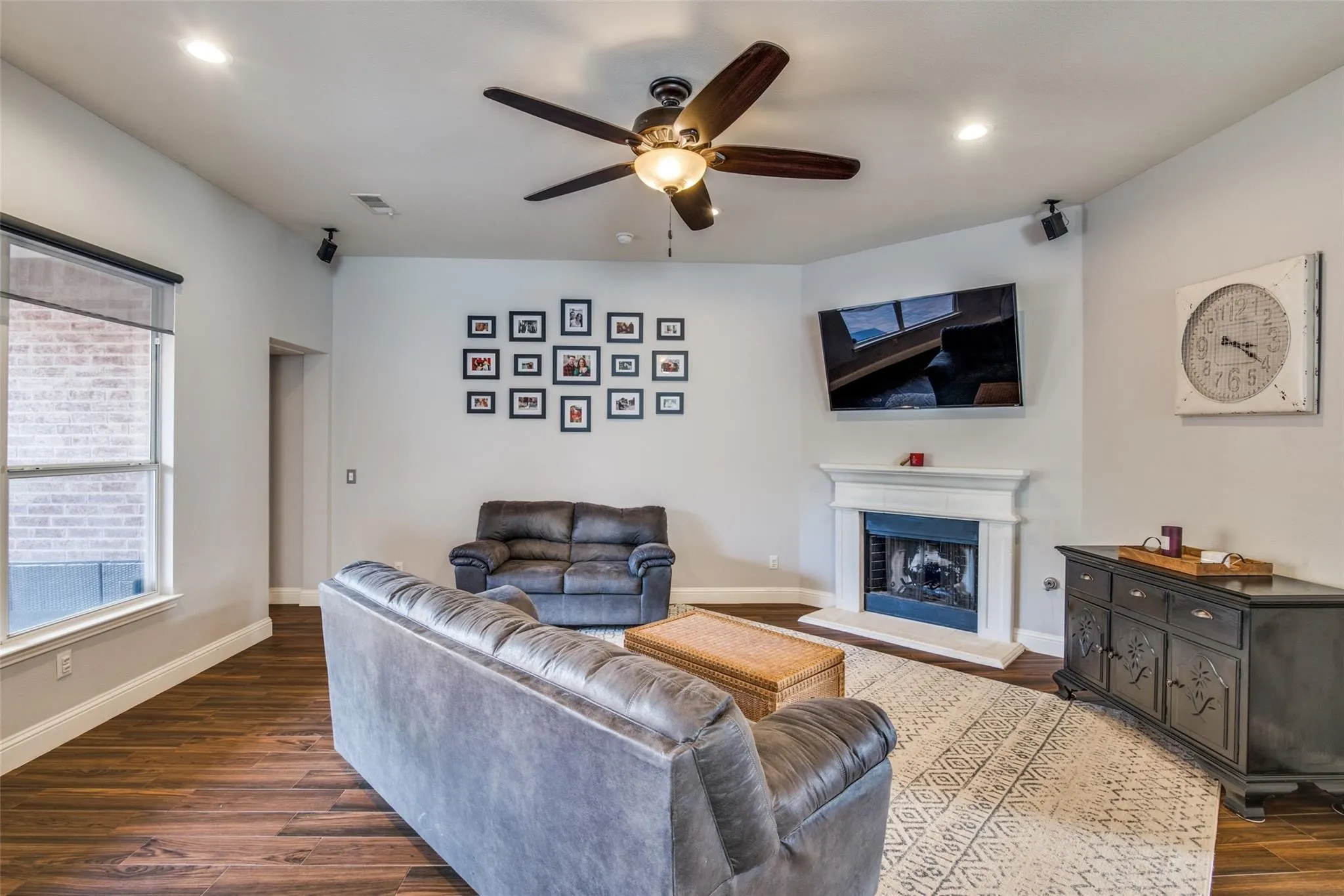 Spacious living area featuring wood-finish flooring, a white mantel fireplace, a ceiling fan with integrated lighting, recessed lighting, and large windows with roller shades