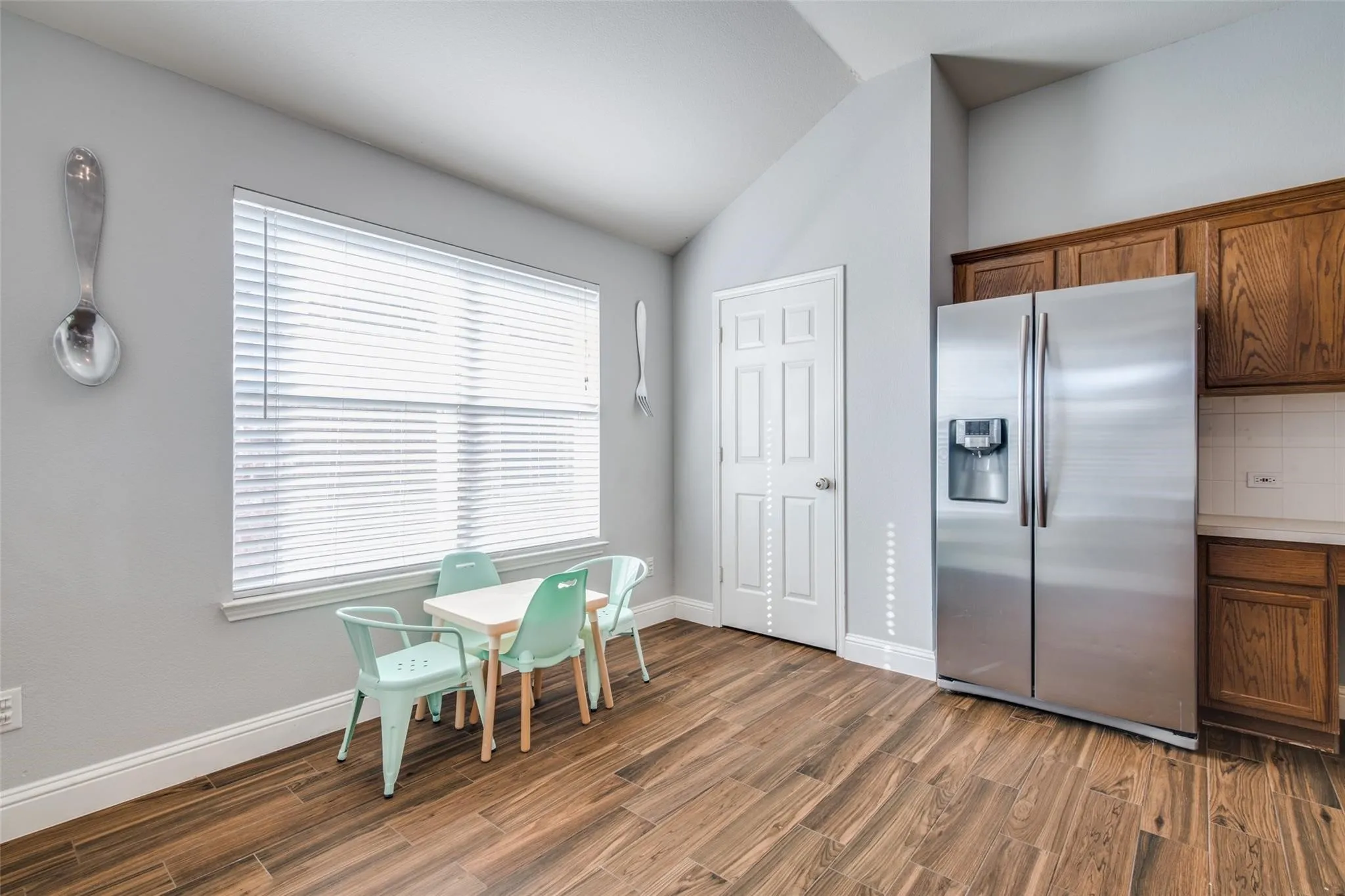 Kitchen area featuring wood-finish flooring, a large window with blinds, vaulted ceilings, a stainless steel refrigerator, and wood cabinetry