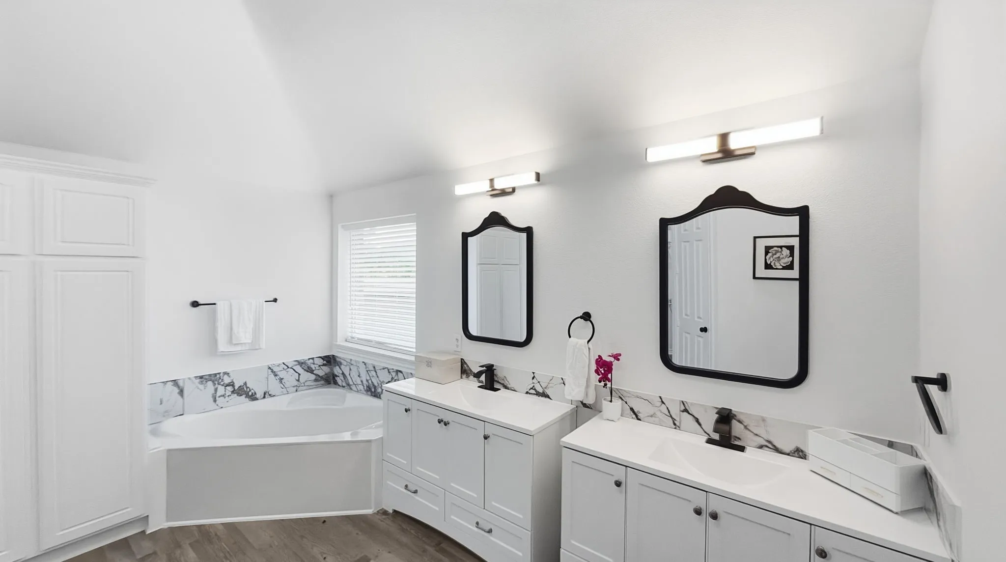White shaker cabinetry, dual vanities with integrated sinks, corner soaking tub, white and grey marble-finish tile, and wood-finish flooring