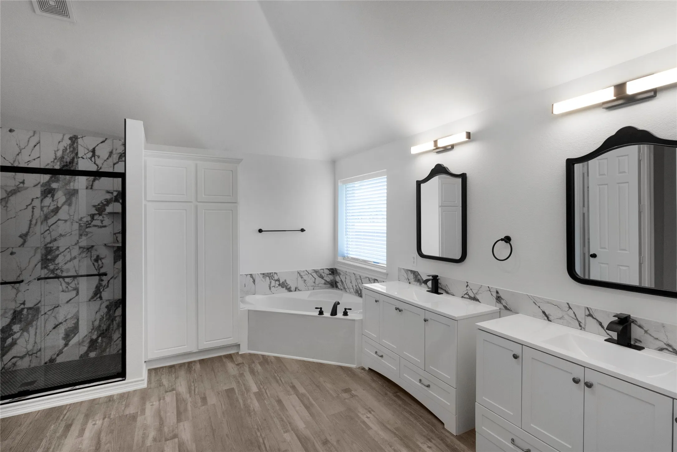 Spacious bathroom featuring a double vanity with white cabinetry, LVP flooring, a built-in soaking tub, a stand-up shower with black-framed glass enclosure, and white marble-look tile accents