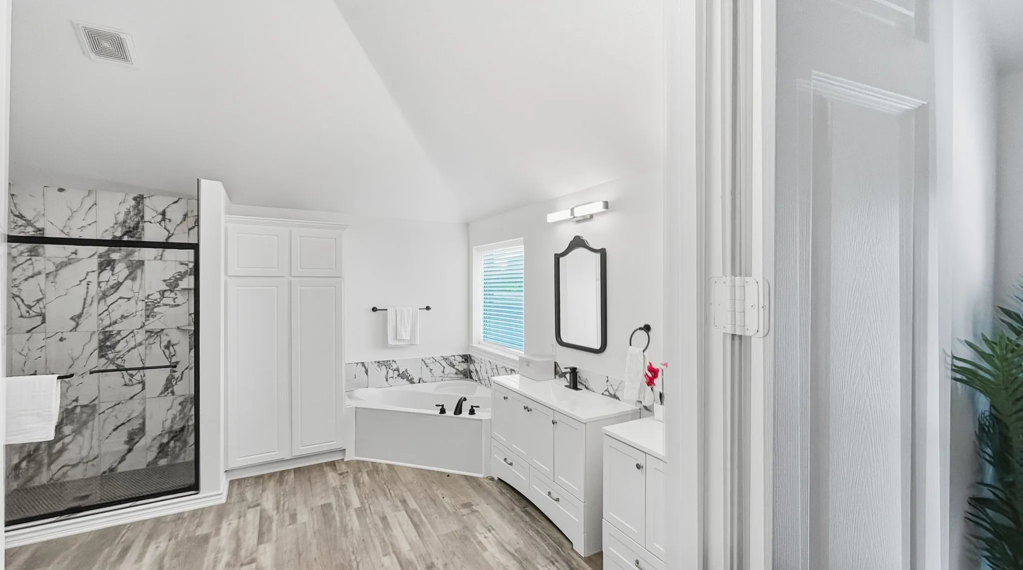 Spacious bathroom featuring a glass-enclosed shower with black-veined tile, a built-in soaking tub, wood-finish flooring, white cabinetry, and a dual vanity