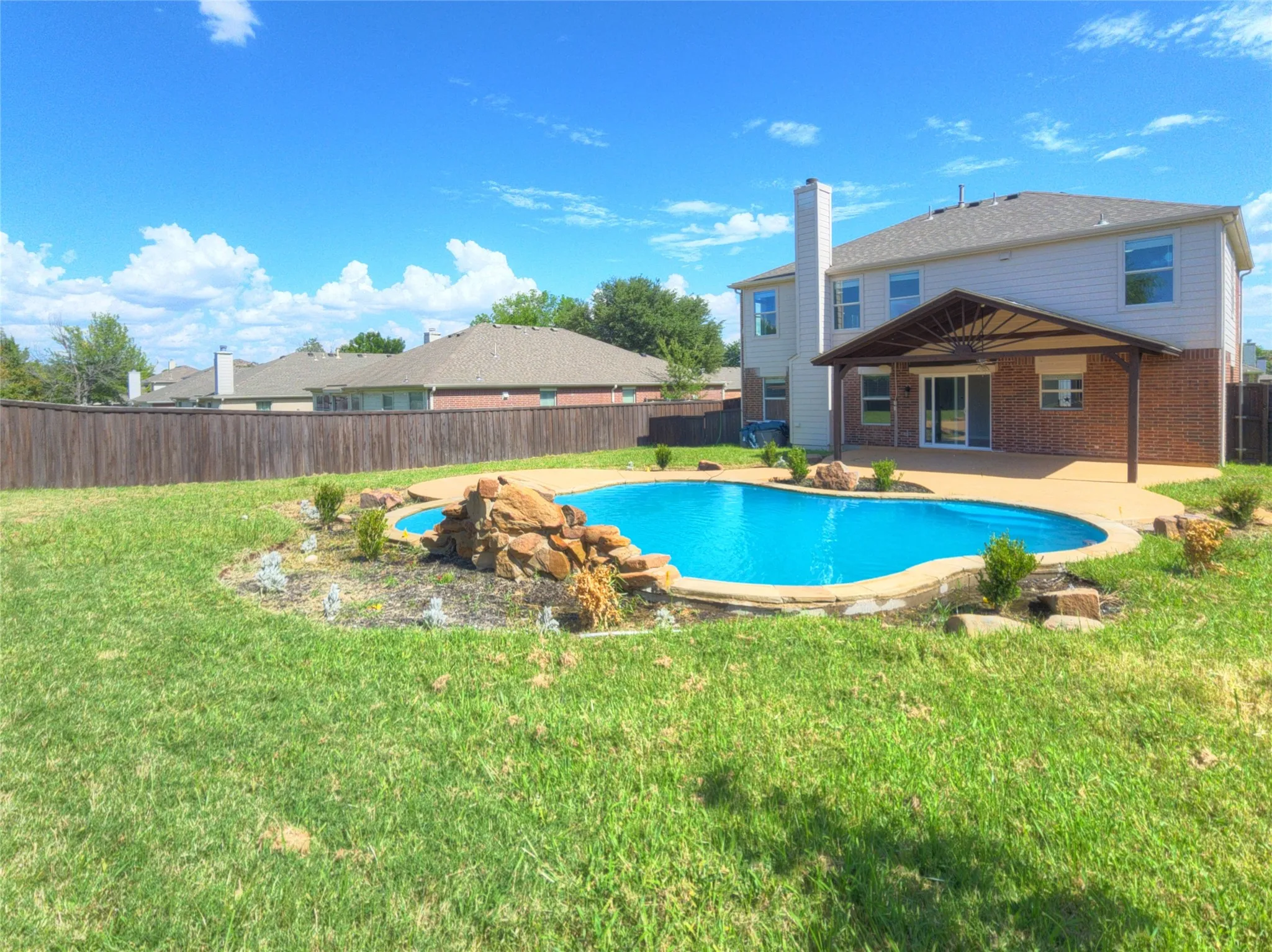 Expansive backyard featuring a blue swimming pool with a rock water feature, a covered patio with an outdoor ceiling fan, a privacy fence, and a two-story home exterior with brick and siding