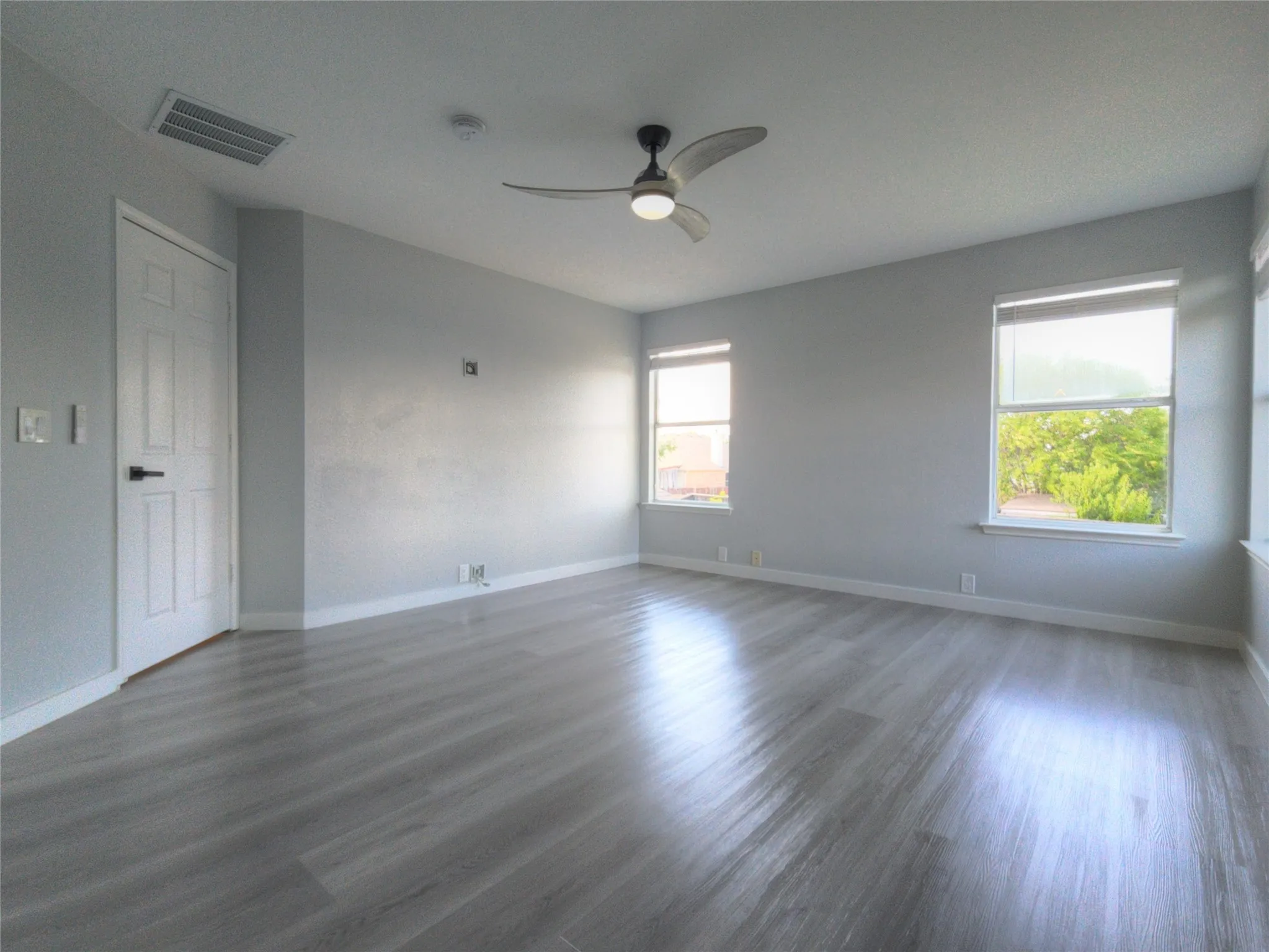 Spacious room featuring light gray walls, wood-finish flooring, and multiple windows