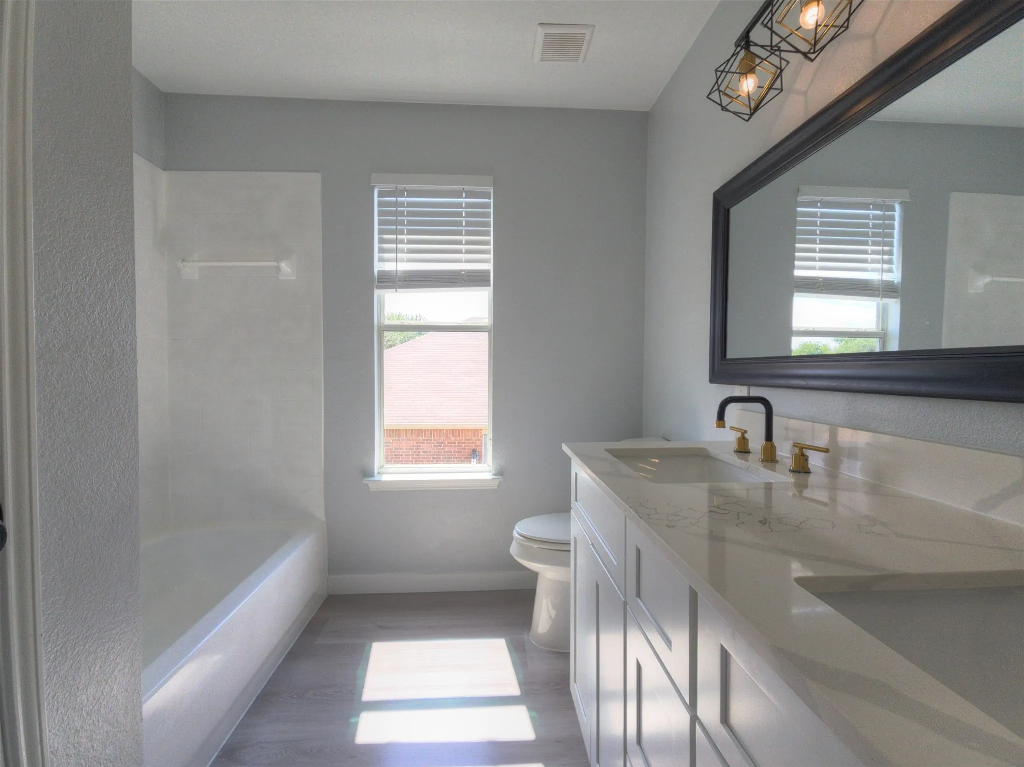 Bathroom featuring a white tub, light wood-finish flooring, a window with blinds, a double vanity with white cabinetry, and a black-framed mirror