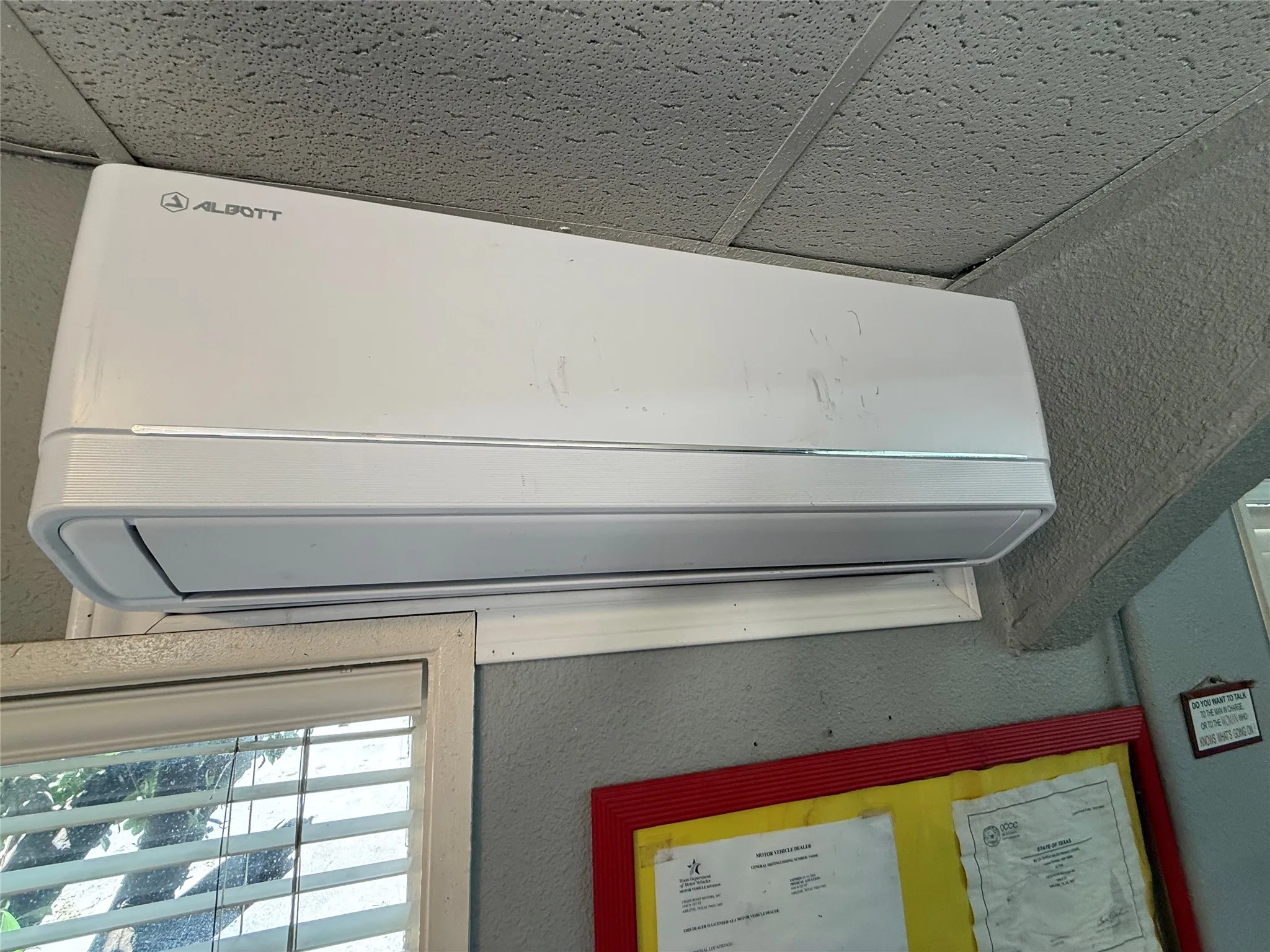 Wall-mounted air conditioning unit positioned above a window with horizontal blinds