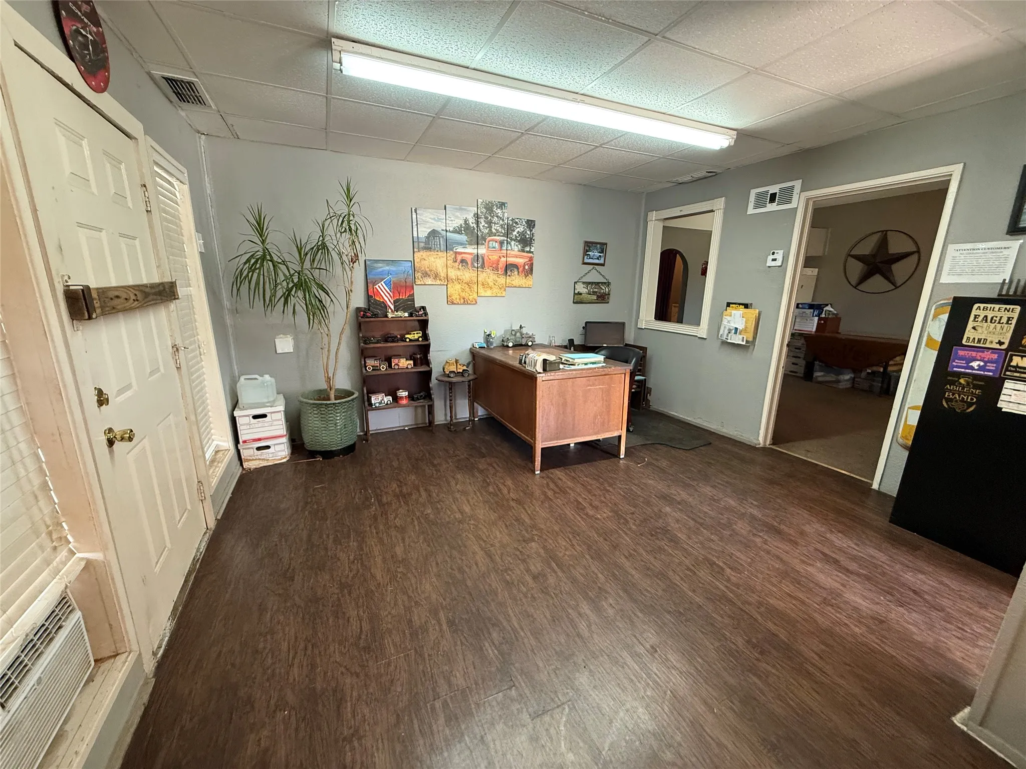 Spacious room featuring wood-finish flooring, drop ceiling with fluorescent lighting, and a paneled entry door