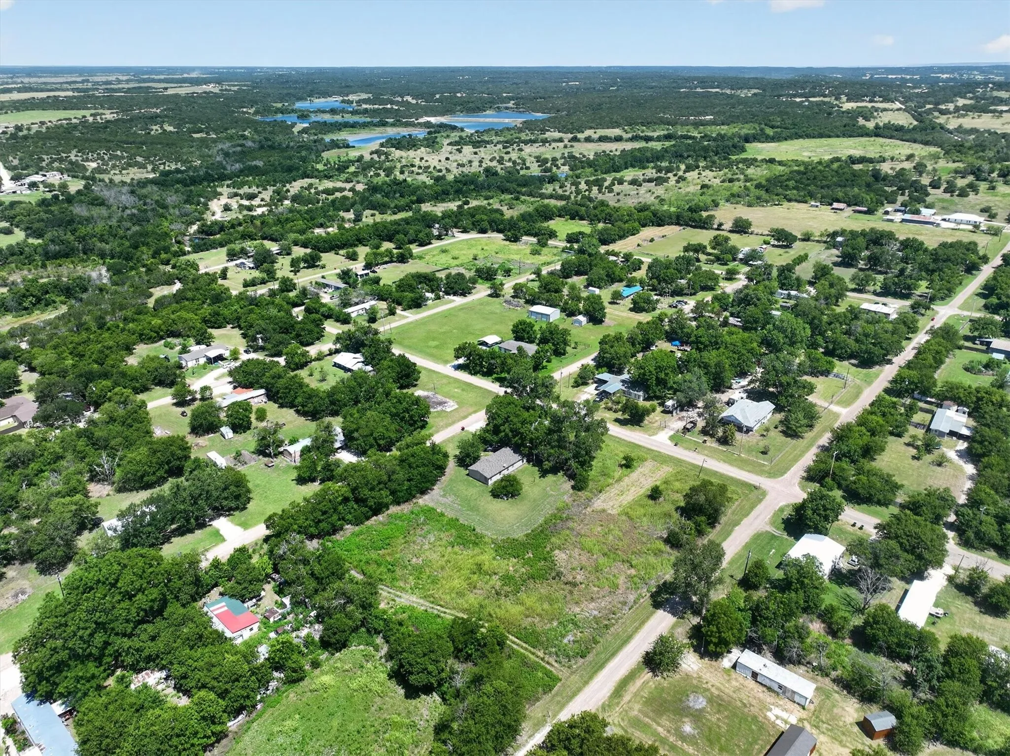 Expansive aerial view of a rural landscape featuring multiple properties, abundant mature trees, open fields, and distant bodies of water