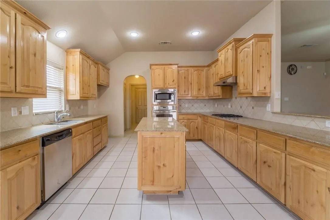 Spacious kitchen featuring a central island, light wood cabinetry, granite-finish countertops, stainless steel appliances, and white tile flooring