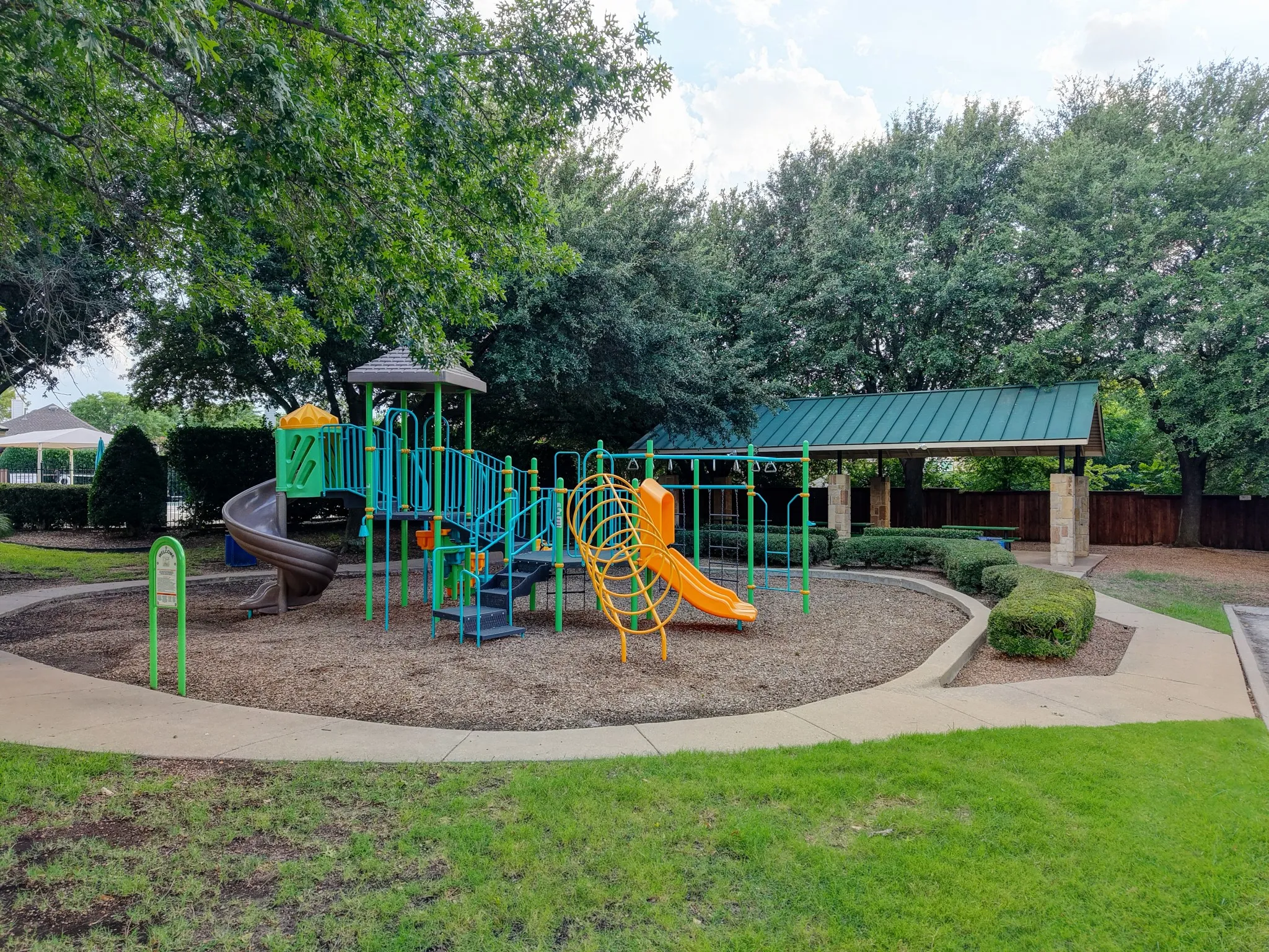 Recreational area featuring a multi-level play structure with slides and climbing elements, a covered pavilion with a standing seam roof, concrete pathways, and mature trees