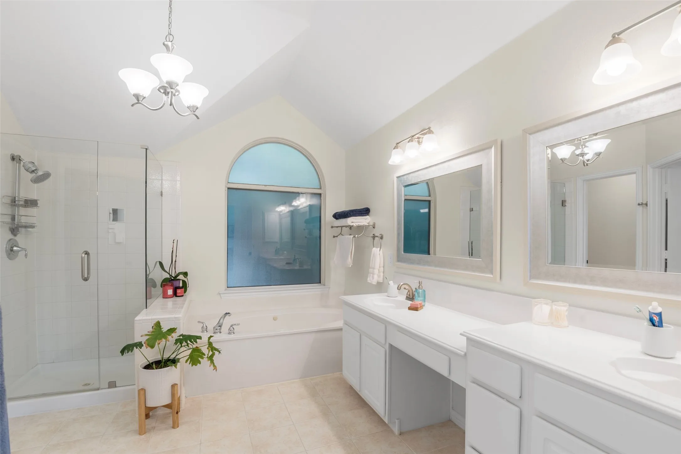 Spacious bathroom featuring a vaulted ceiling with chandelier, an arched window, a built-in jetted tub, a separate glass-enclosed shower, and a double vanity with ample counter space