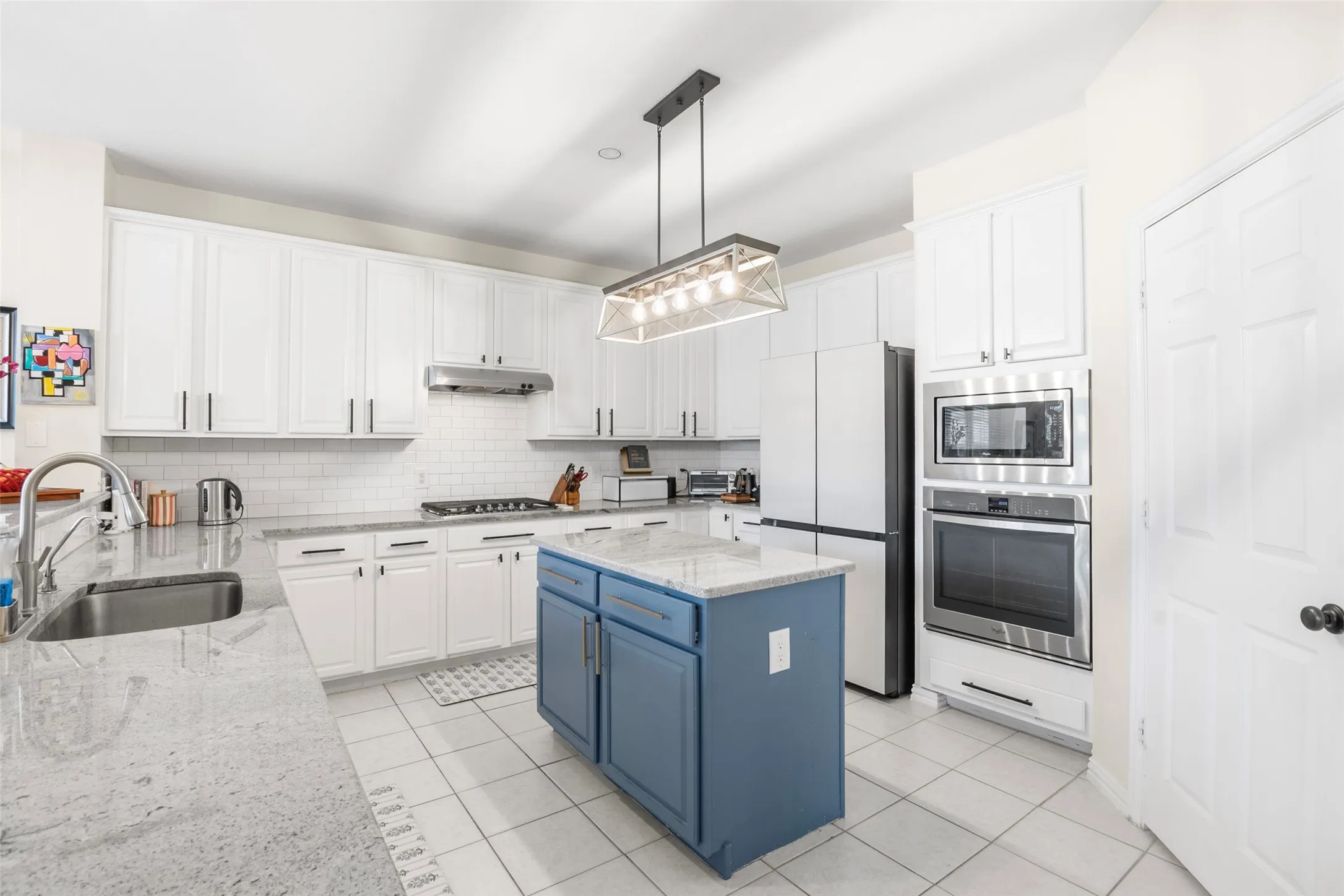 Modern kitchen featuring white cabinetry, light-toned countertops, a blue center island, stainless steel appliances, and white tile flooring