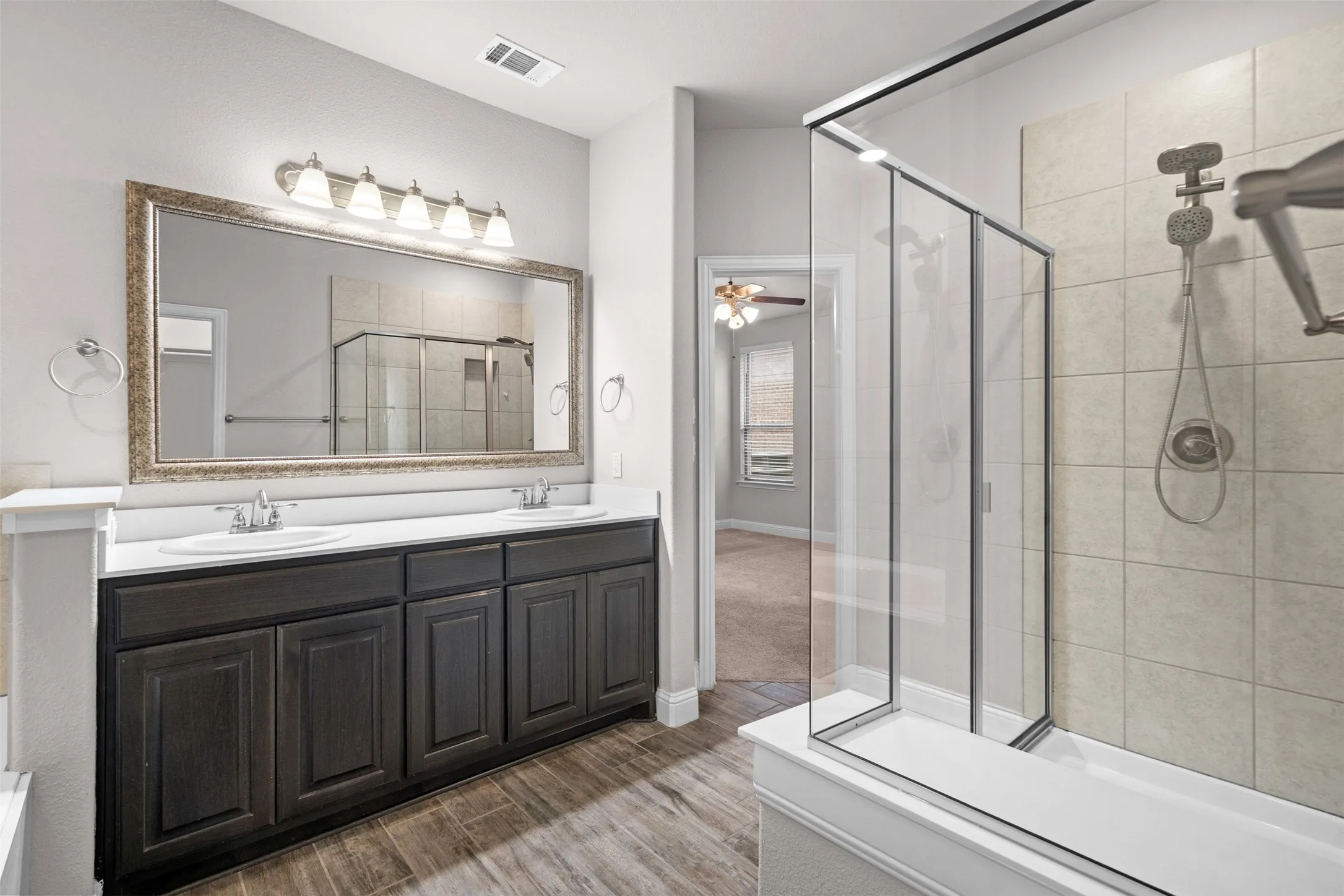 Bathroom featuring a dual vanity with dark wood-finish cabinetry, a large framed mirror, and a glass-enclosed shower with tiled walls and a handheld shower head