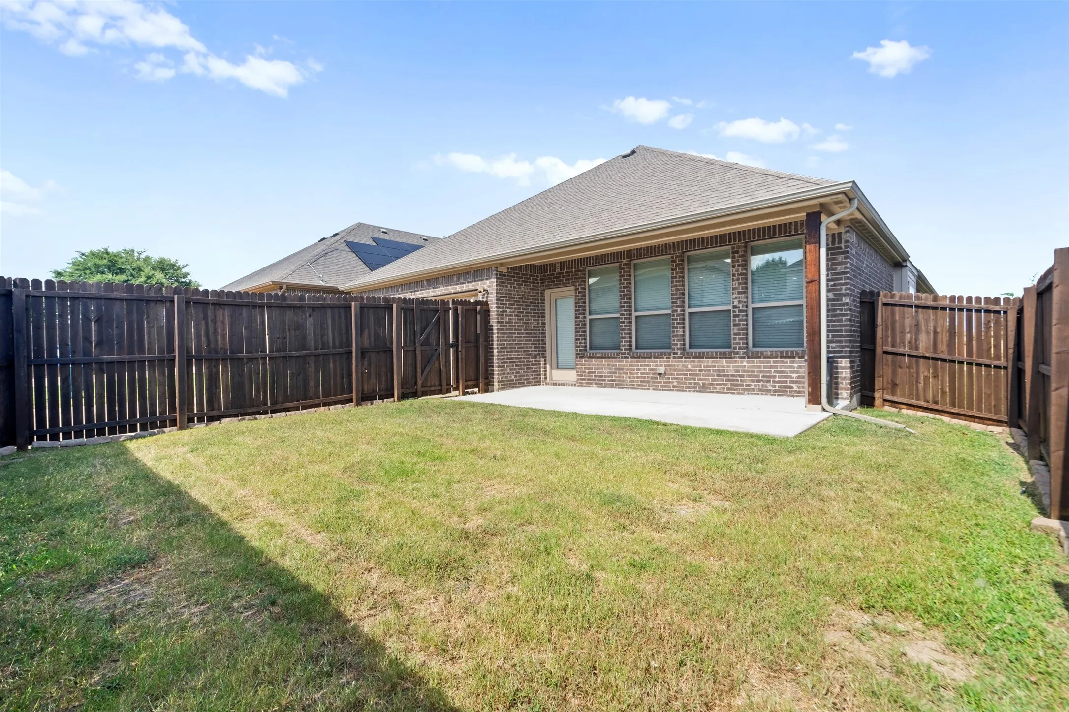 Expansive grassy area featuring a concrete patio, full privacy fencing, and a brick exterior with multiple windows