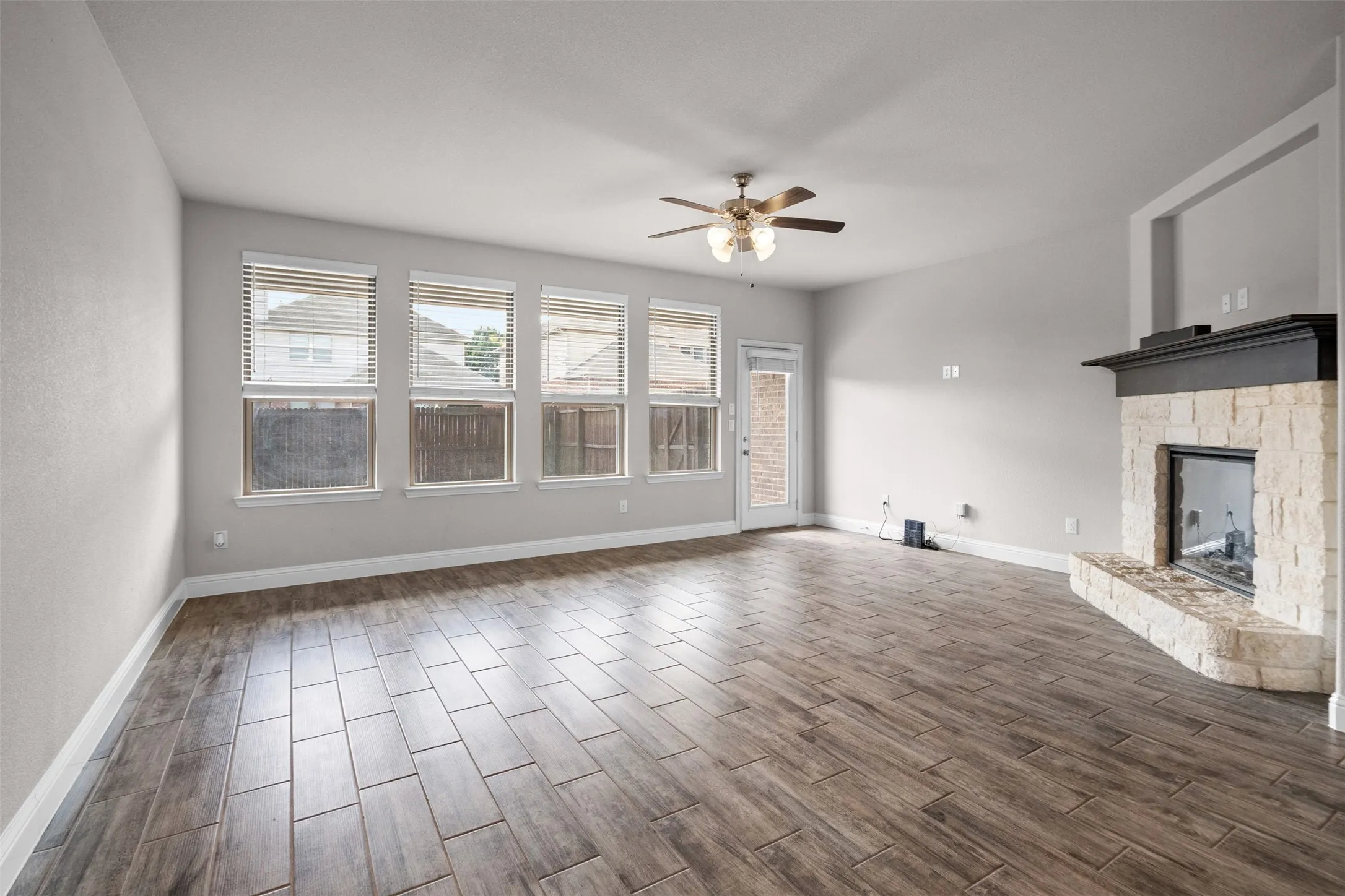 Spacious living area featuring wood-finish flooring, a stone-clad fireplace with a dark mantel, and multiple windows with blinds