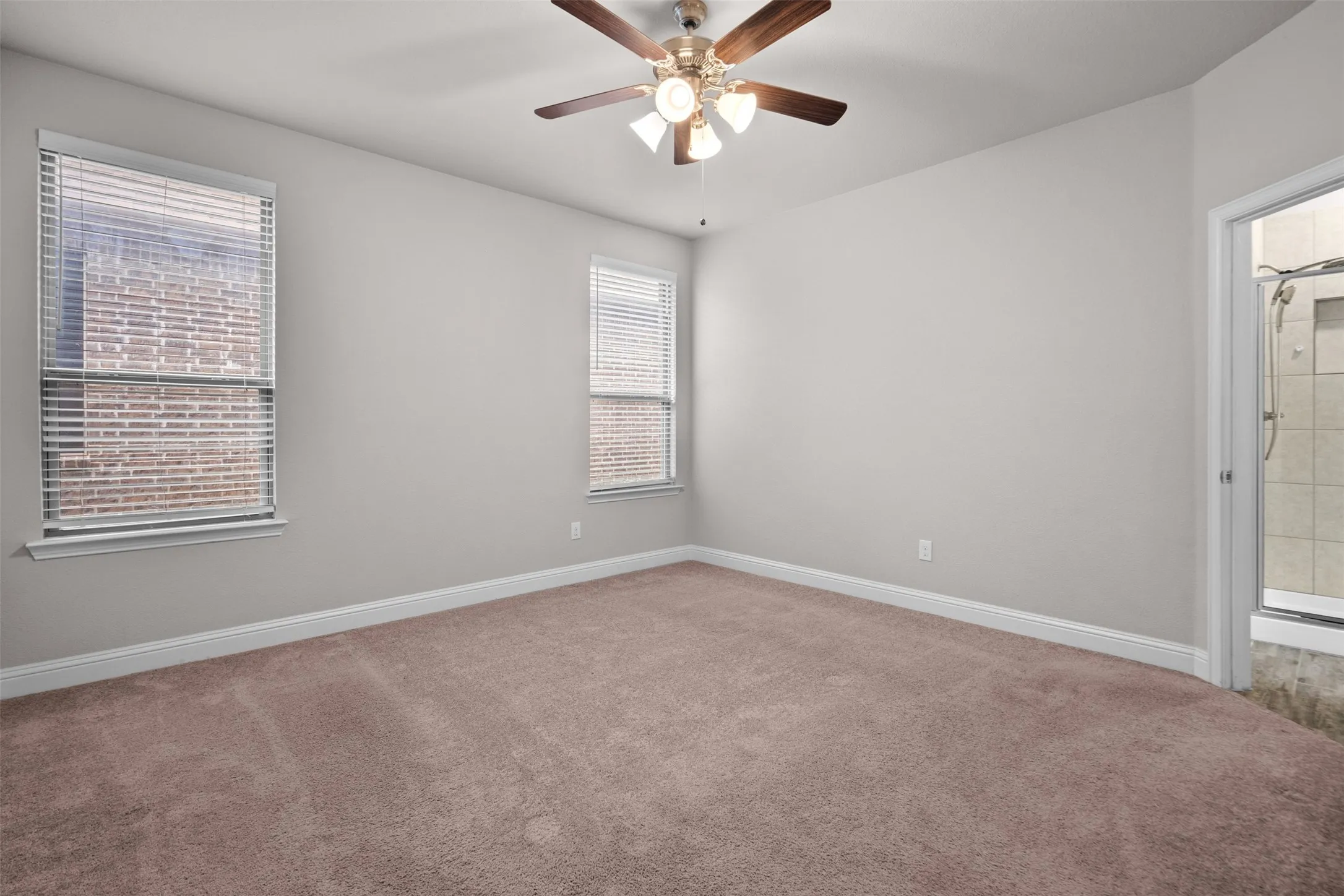Carpeted room featuring neutral wall paint, two windows with blinds, a ceiling fan with integrated lighting, and direct access to a tiled shower enclosure