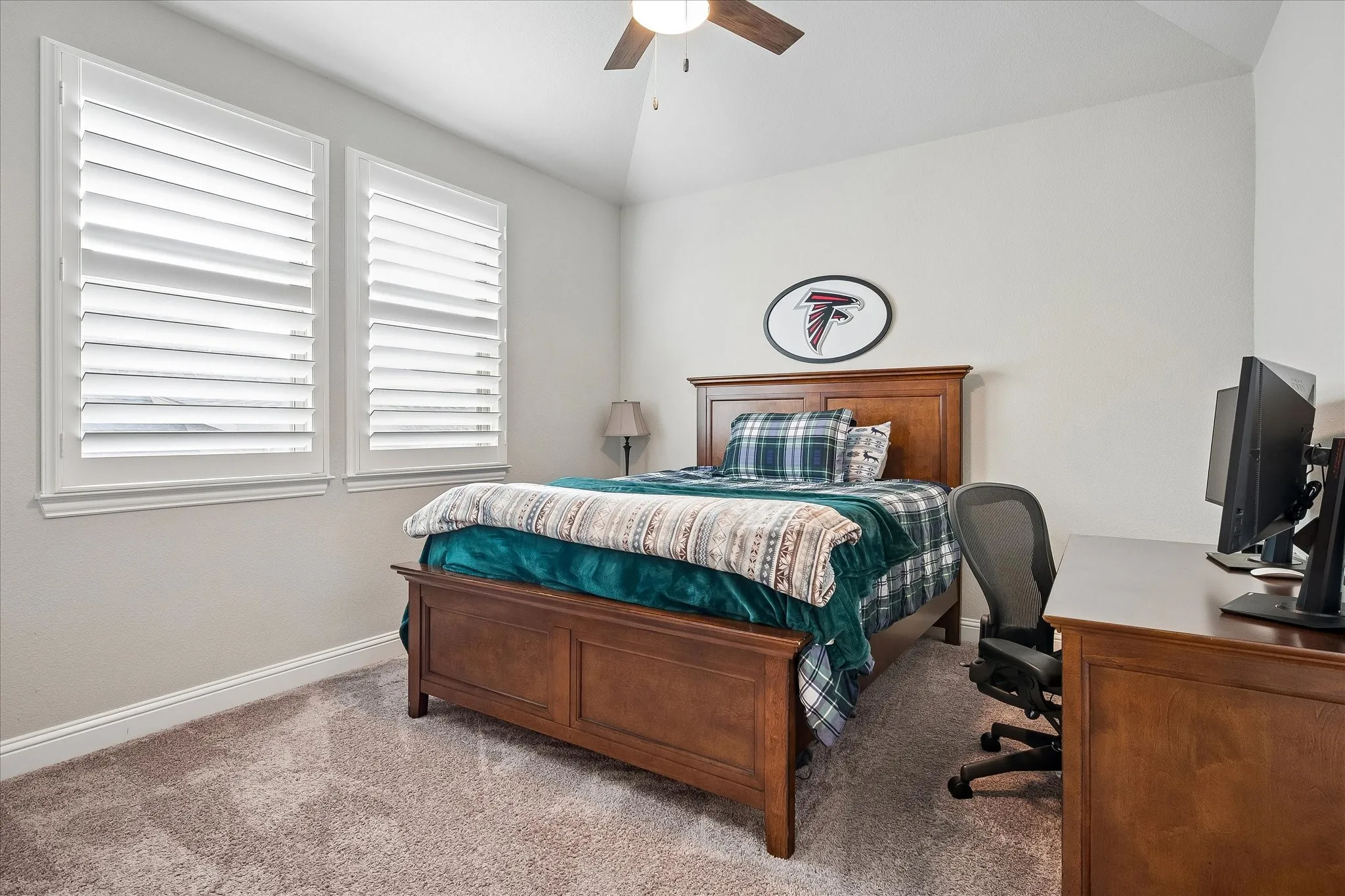 Comfortable room featuring two windows with plantation shutters, plush carpeting, a wood-finish bed frame, and a wood-finish desk