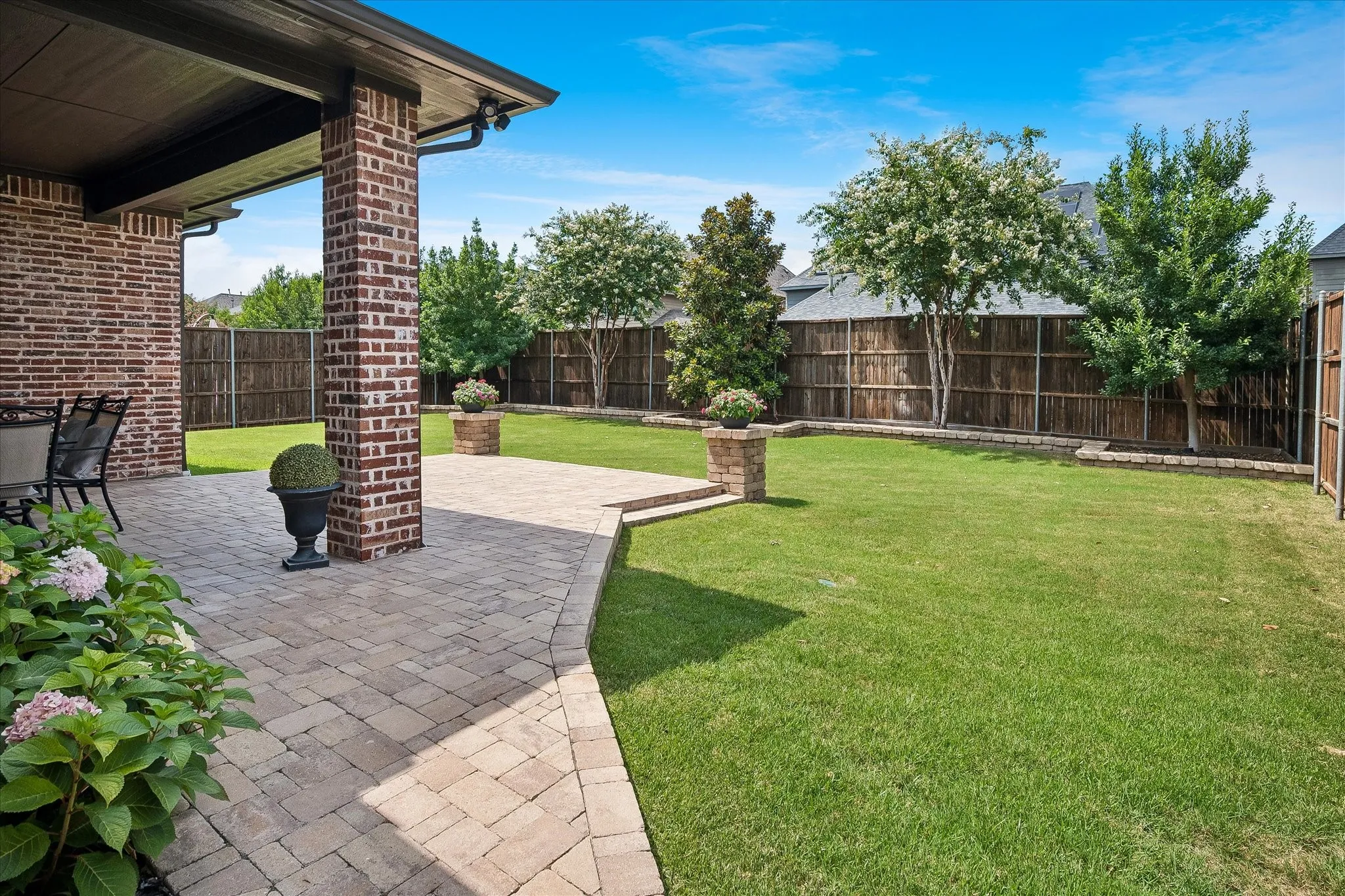 Expansive backyard featuring a covered patio with brick columns, paver stone hardscaping, a manicured lawn, and mature perimeter trees