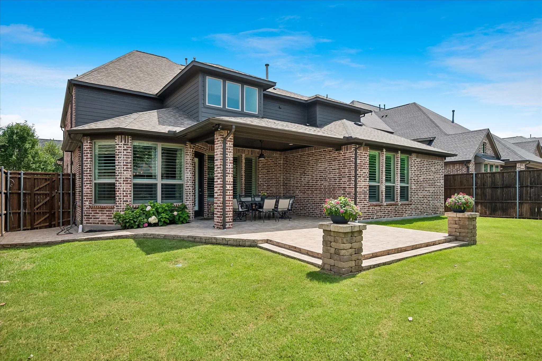 Expansive backyard featuring a brick exterior with dark siding, a covered patio area, a stepped paver patio, a manicured lawn, and privacy fencing