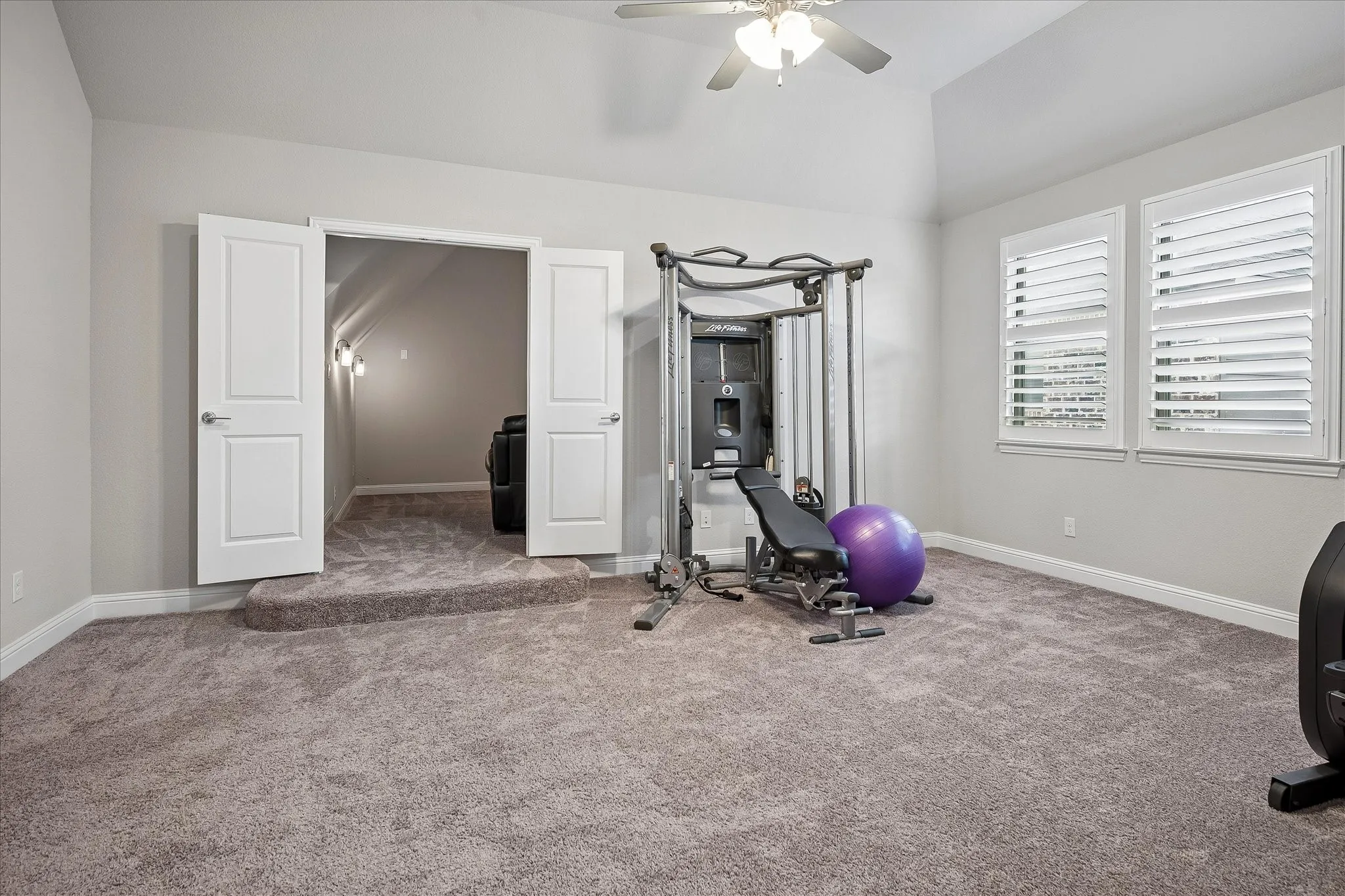 Spacious carpeted room featuring vaulted ceilings, a ceiling fan with integrated lighting, and two windows with white plantation shutters