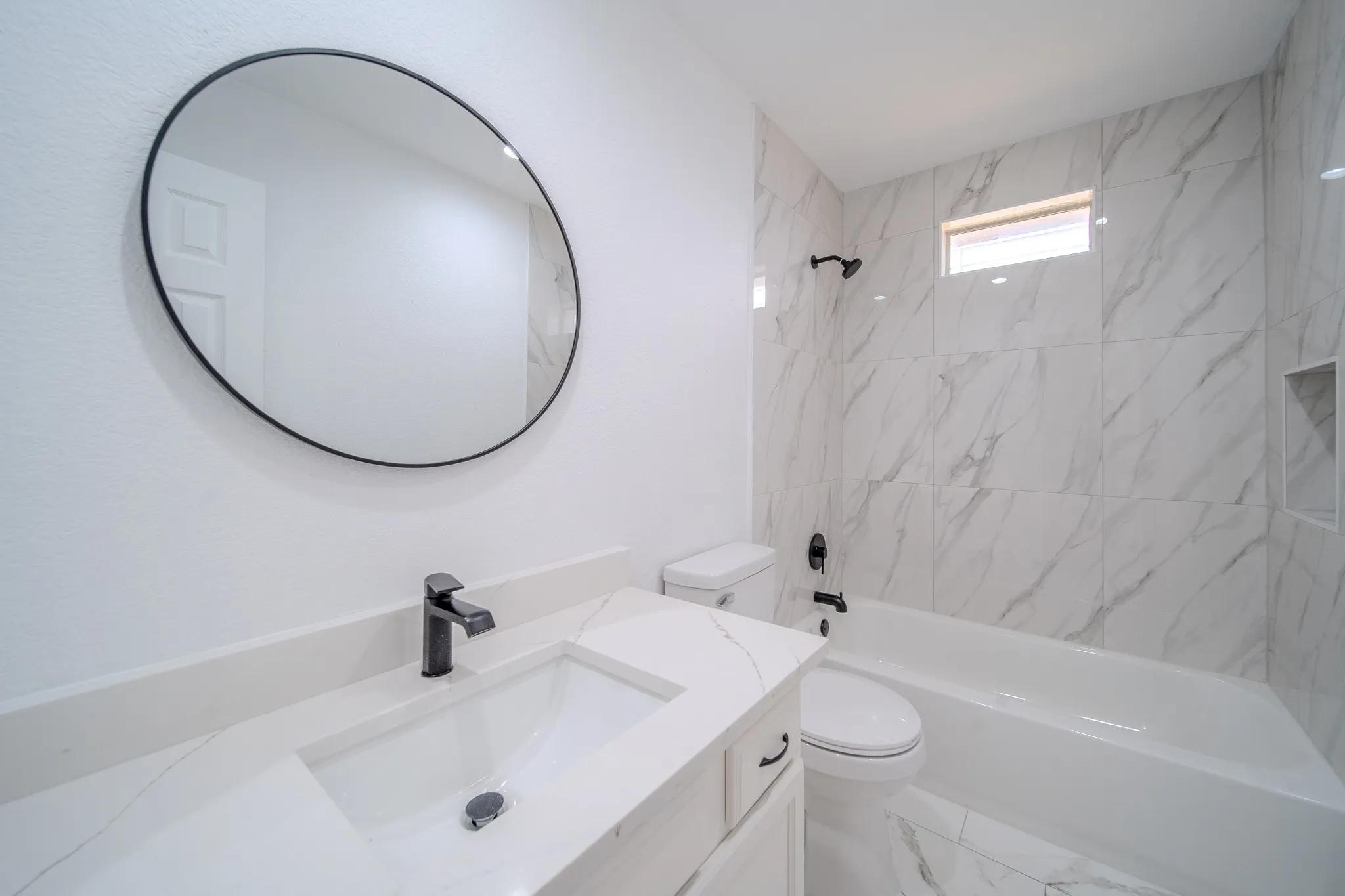 Contemporary bathroom featuring a white vanity with an integrated sink, a large circular mirror with a black frame, black fixtures, and marbled wall and floor tiling